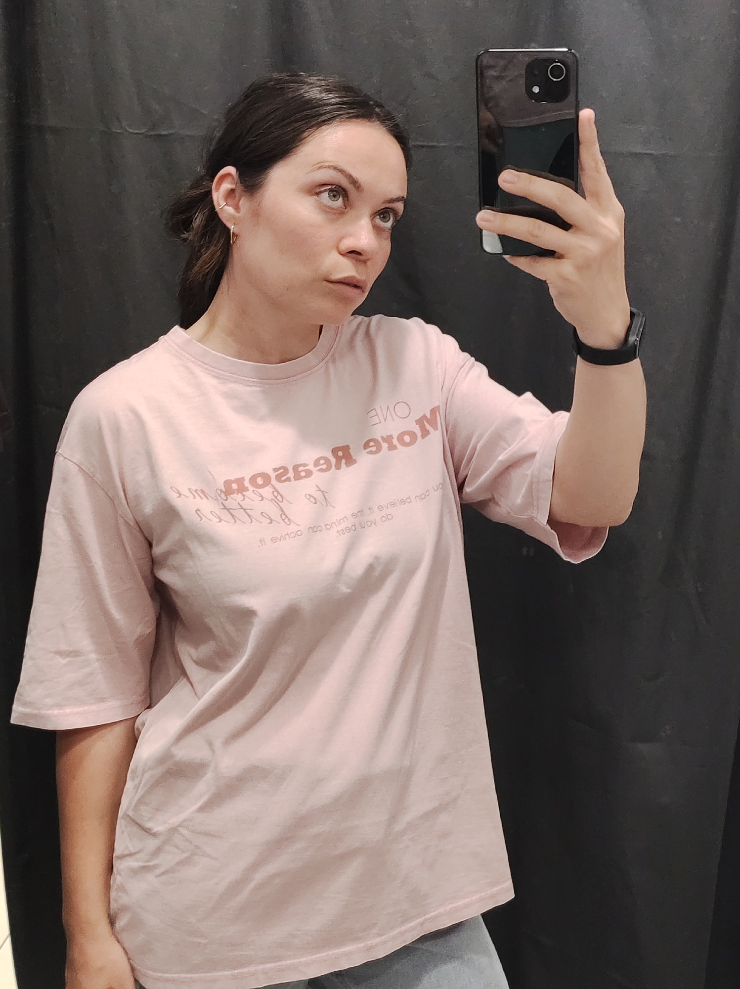 A young woman is taking a selfie in what appears to be a changing room or small space with a dark gray curtain in the background. She is wearing a light pink oversized t-shirt with a printed message on the front. The text on the t-shirt is partially reflected and distorted due to the angle, but some words are legible such as "ONE MORE Reason." She is holding a black smartphone in her right hand to capture the photo. She has dark hair pulled back in a casual style and is wearing a black fitness tracker on her wrist and a small earring. The image does not offer any details of the city of Chaikovsky, Russia.