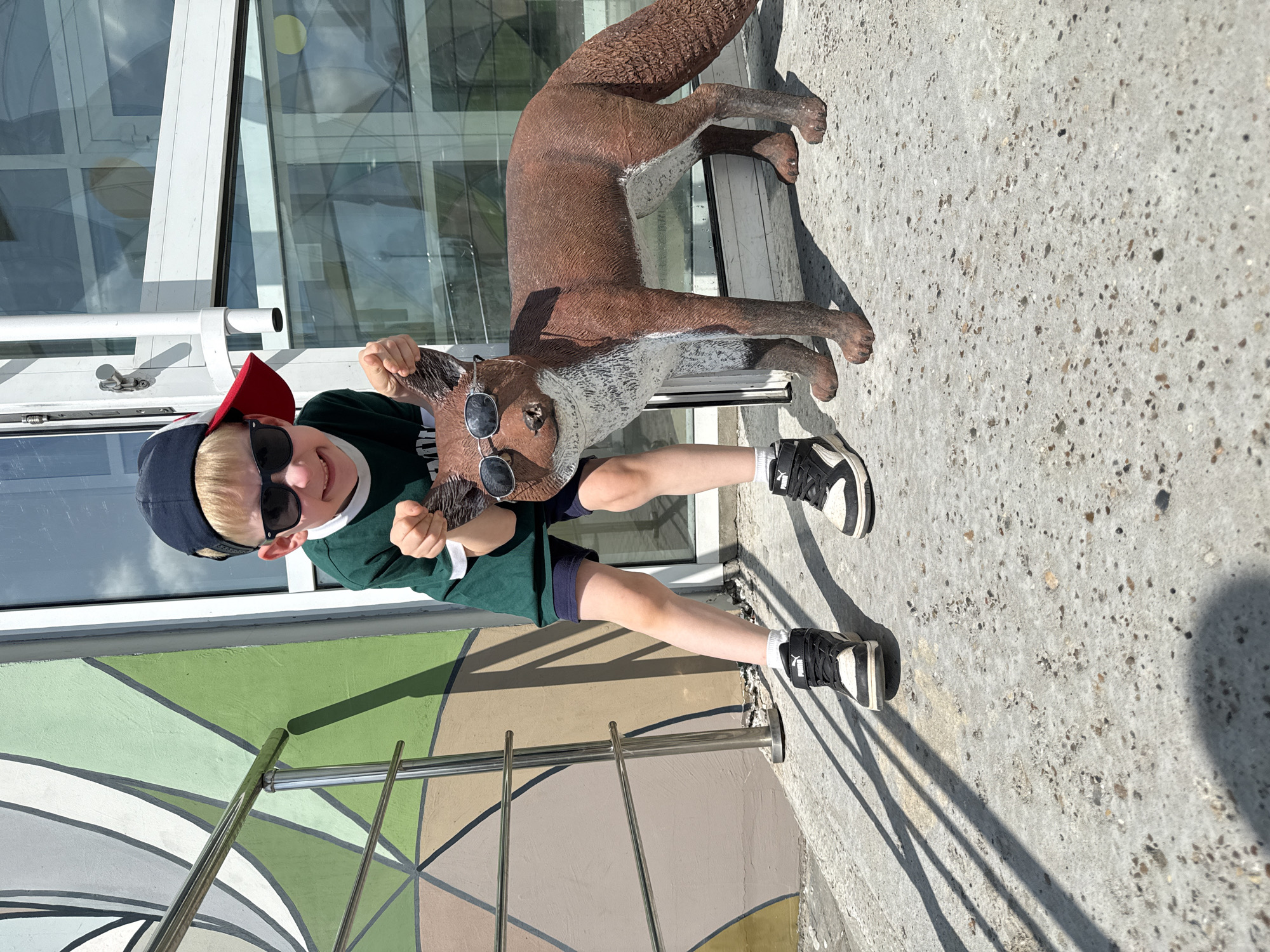 A young boy, wearing sunglasses and a red and navy baseball cap, stands next to a life-sized statue of a fox wearing sunglasses. The boy is wearing a green t-shirt, dark shorts, and black and white sneakers. He is holding the fox statue by its ears, with his mouth open in a grimace. The fox statue is brown and white, with a bushy tail. They are standing on a concrete sidewalk in front of a building with large windows. A mural with geometric shapes is visible on the wall to the left. The scene appears to be outdoors on a sunny day. The location is identified as Perm, Russia.