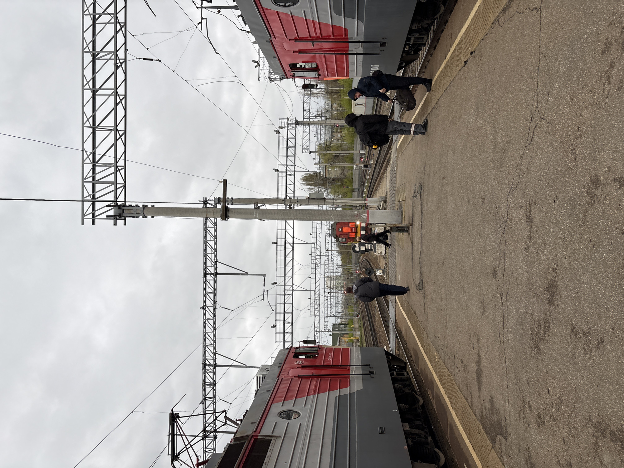 The image shows a train station platform in Saratov, Russia, flanked by two gray and red locomotives. Overhead, a network of steel support structures and electrical lines dominates the sky.
On the platform, two individuals are near one of the trains, and another individual is standing near the opposite train. The sky is overcast and gray. The platform is made of asphalt with a yellow tactile strip near the train tracks.