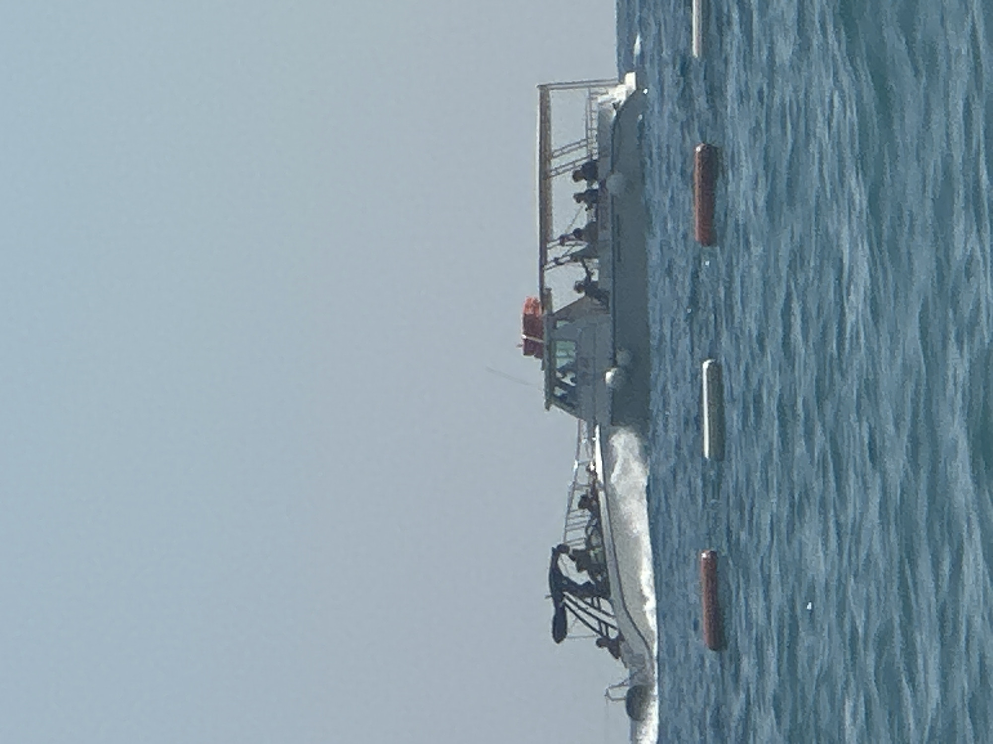 Two boats are navigating through the choppy blue waters of the sea. The boat on the left is a smaller speed boat with a dark canopy, carrying several individuals. It is creating a spray of water as it moves forward. The boat on the right is larger, a passenger boat with a white hull and a tan canopy, also carrying a group of people. In the foreground, three buoys are floating on the water: one is dark red and the other two are light colored. The sky above is clear and light blue, suggesting fair weather. The scene appears to be taking place off the coast of Boreti, Montenegro.
