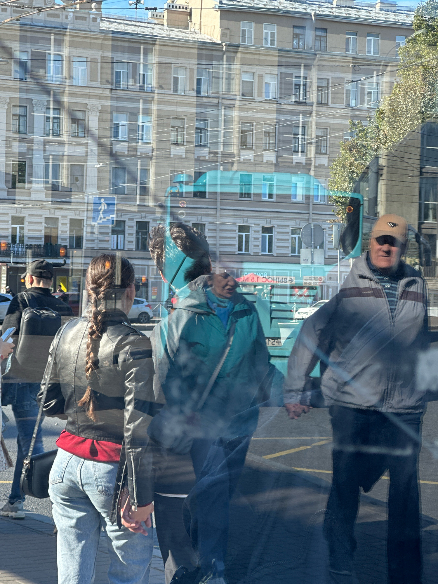 This is a reflection of a street scene in Saint Petersburg, Russia, on a window. The scene is superimposed with the reflection of a building.
In the foreground, a young woman with her back to the camera is wearing a black leather jacket, a red top, and light blue jeans. Her hair is in a braid. She is holding a black purse with a strap over her shoulder. Next to her, a man in a tan baseball cap and a gray jacket is visible, holding hands with a person wearing a teal jacket, whose face is partially obscured by a reflection. Further back, a person wearing a black cap and black jacket is walking away from the camera.
The background reflection shows a multi-story building with numerous windows. There are also reflections of a turquoise-colored bus or tram, a pedestrian crossing sign, and traffic lights. The environment appears to be an urban street. The lighting suggests it is daytime.