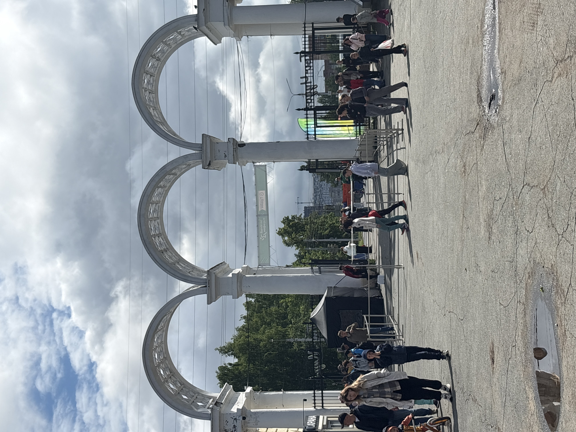 The image captures a street scene at the entrance to Gorky Park in Izhevsk, Russia. An architectural structure of three arches supported by white columns defines the entrance. People of various ages and dress are walking in both directions through the entrance. A small black tent-like structure stands to the right of the entrance. Metal fencing lines a portion of the entrance. In the background, trees and buildings are visible, with overcast skies above. A puddle on the pavement reflects part of the scene.