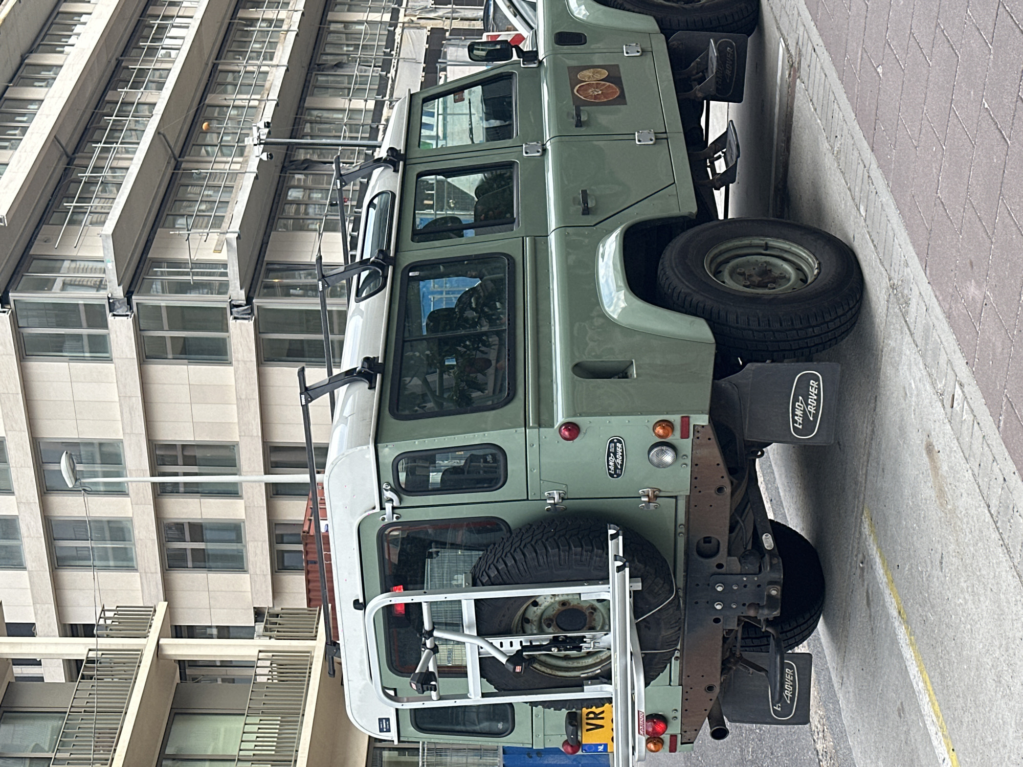 A light green Land Rover Defender is parked on a paved street in Amsterdam, Netherlands. The vehicle features a white roof and a spare tire mounted on the rear. The building behind the vehicle has multiple windows. There is a lamp visible in the background.