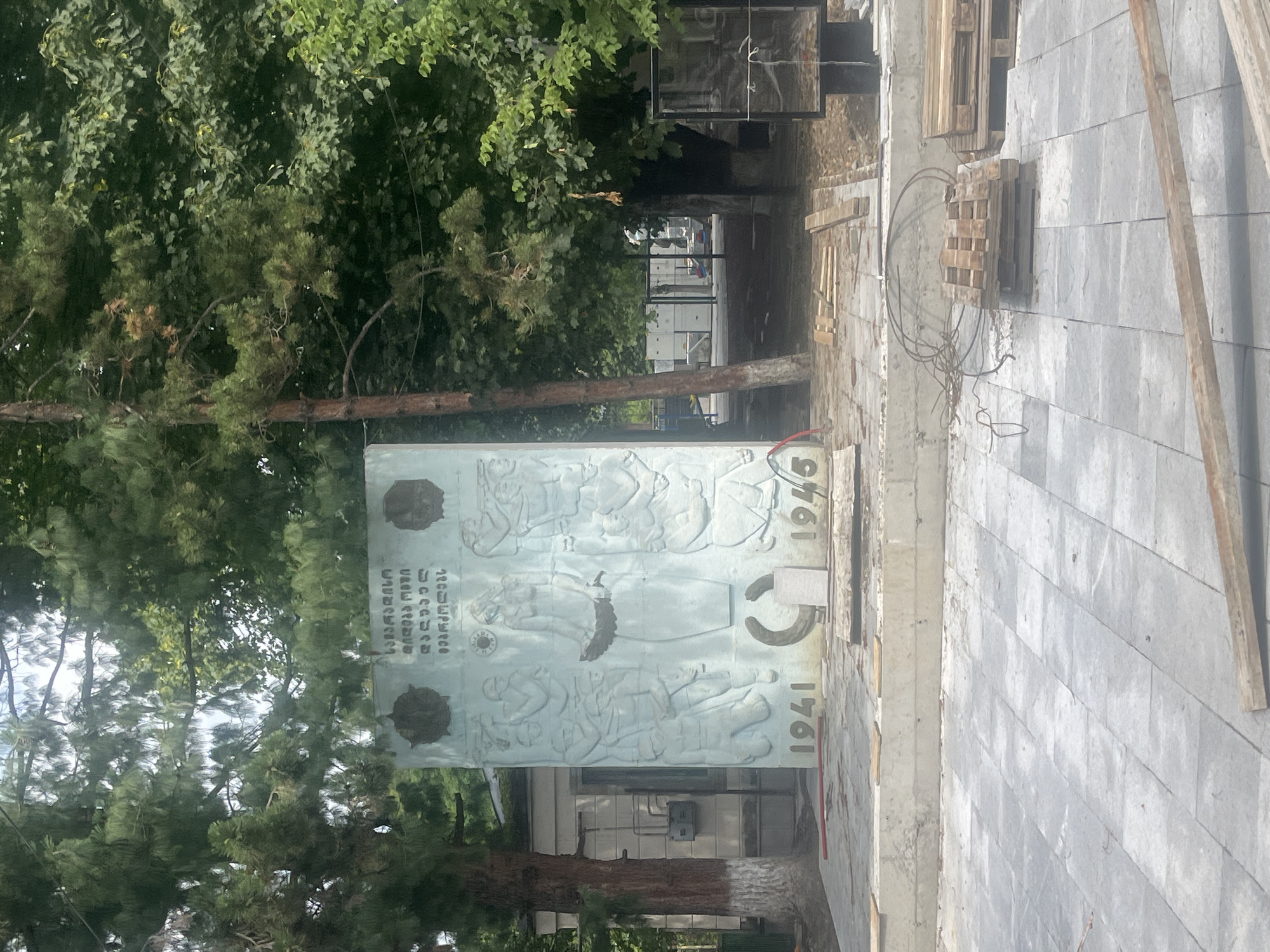 In Surami, Georgia, a memorial monument with relief carvings stands partially obscured by trees. The monument features a central figure of a woman holding a wreath and scenes of soldiers in battle. The years "1941" and "1945" are prominently displayed at the base of the monument. The surrounding area appears to be undergoing construction or renovation, with stacks of wooden pallets and construction materials present on the paved ground. A building with gray siding is visible behind the monument to the left. The sky is overcast.