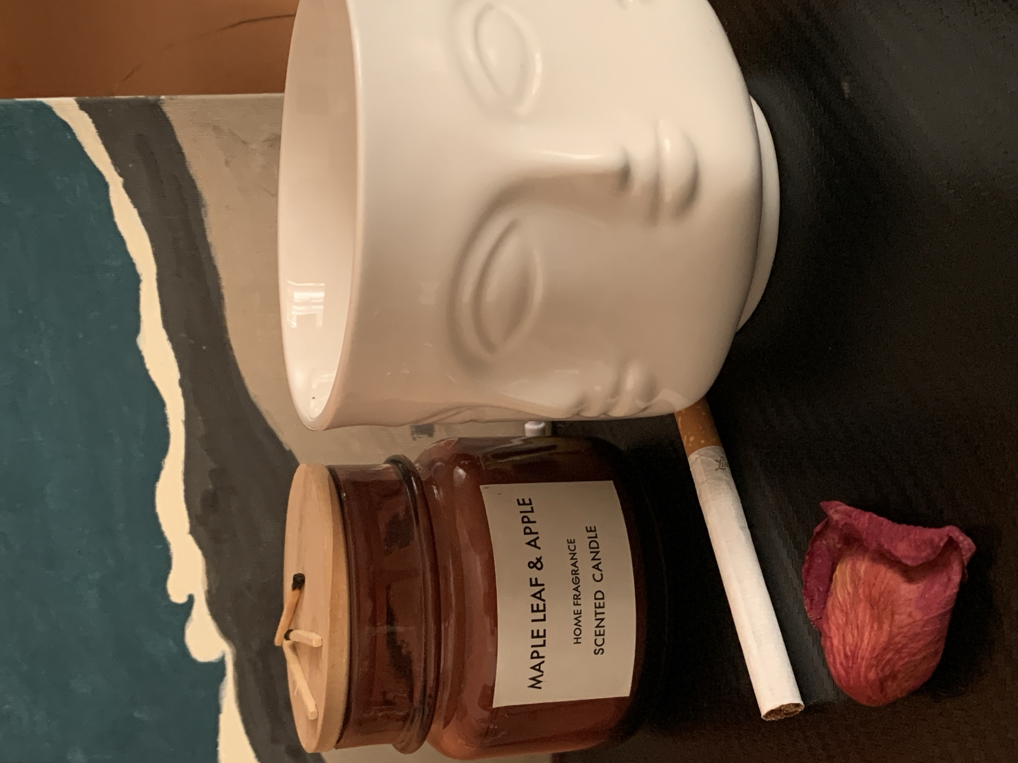 A still-life composition including a white ceramic vase molded to resemble a human face; a brown candle labeled "Maple Leaf & Apple," with matches lying on the wooden lid; a single cigarette; and a single rose petal. The scene appears to be indoors. The items are arranged on a dark surface. The vase is positioned at the top, and the candle, cigarette, and rose petal are at the bottom. The location is in the city of Bar, Montenegro.