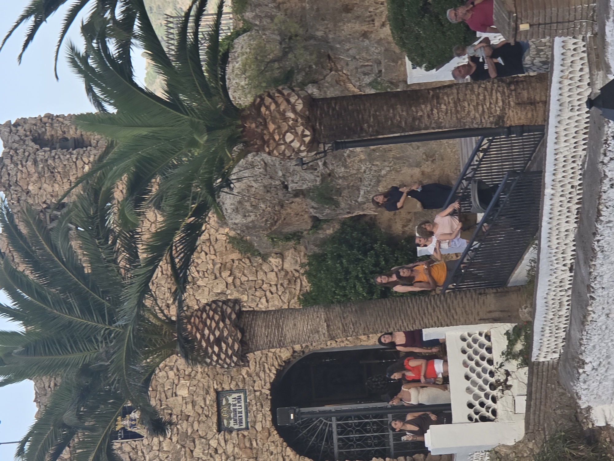 The image depicts the exterior of the Santuario de la Virgen de la Peña in Mijas, Spain. The sanctuary, constructed with rough-hewn stones, features an arched entryway with an ornate metal gate. Two prominent palm trees stand in front of the structure, their fronds casting shadows. Several people are visible on a white railing and a black staircase leading up to the sanctuary.

On the left side, near the entrance, a group of women stands, some looking towards the camera. One woman in a white dress and another in a red top are particularly noticeable. Further to the right, a woman in an orange top is visible on the stairs, interacting with a person holding a baby. To the far right, two men are standing on a lower level, one holding a baby. The environment appears to be outdoors, with rocky terrain and some greenery visible in the background. The lighting suggests it is daytime, possibly late afternoon.