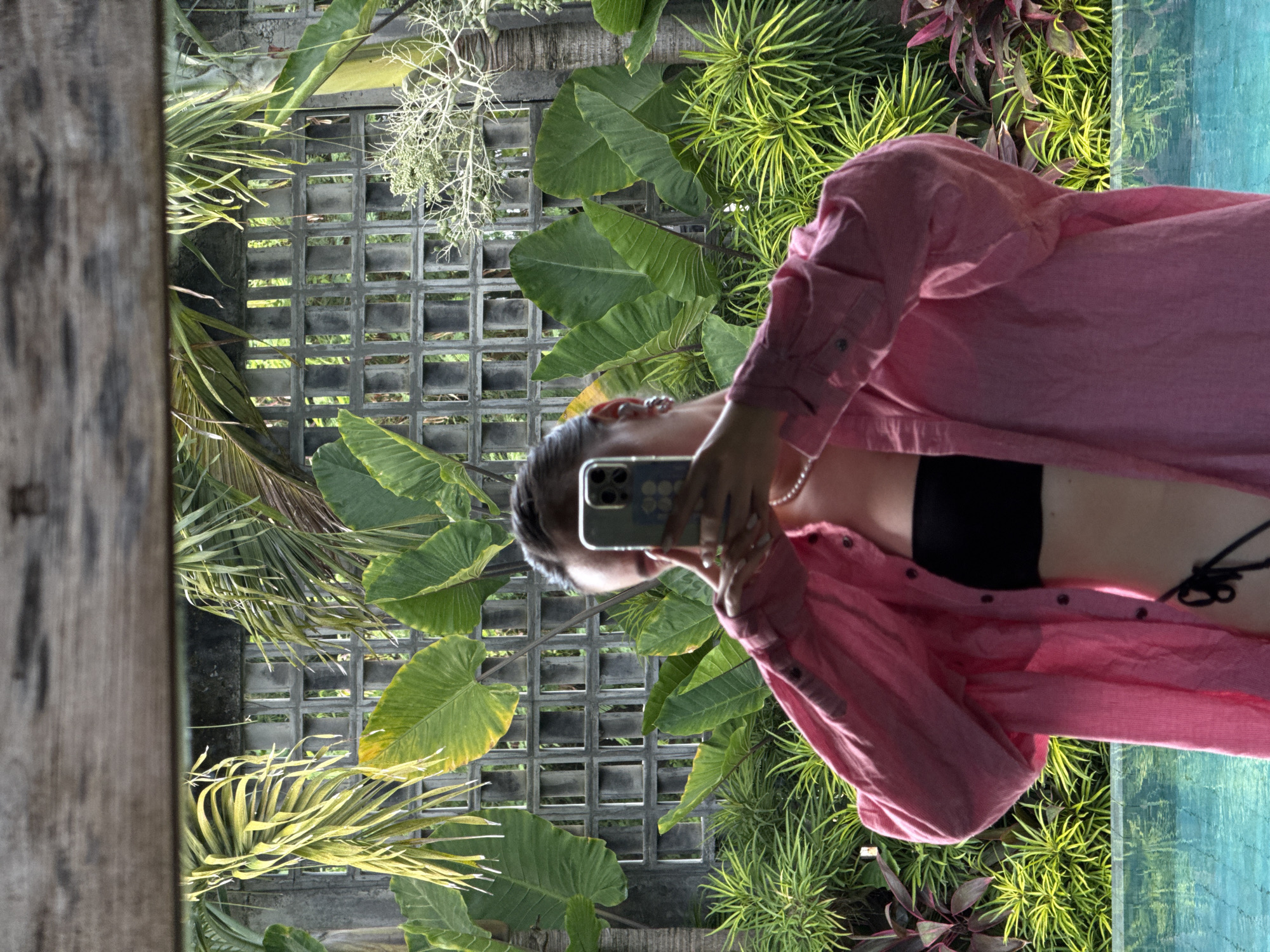 The image, rotated 90 degrees counter-clockwise, depicts a person taking a mirror selfie in an outdoor, tropical-themed setting during bright daylight. The subject, positioned horizontally with her head to the right, is a person (likely female) with dark hair. She is seen wearing a long-sleeved, unbuttoned pink shirt over a black top, possibly a bikini top or bralette. A pearl necklace adorns her neck, and rings are visible on her fingers as she holds up a light-colored smartphone, encased in a pale blue cover, to take the photograph.
The background, visible as a reflection, showcases lush green and reddish-purple tropical foliage. A grey concrete lattice wall with square openings forms a prominent part of the scene, with more greenery peeking through its gaps. To the far right, a section of bright, light blue water, likely a swimming pool, is partially visible. A rustic wooden post or frame runs vertically along the left edge of the image, suggesting the selfie is taken in a mirror or reflective window. No discernible text is visible.