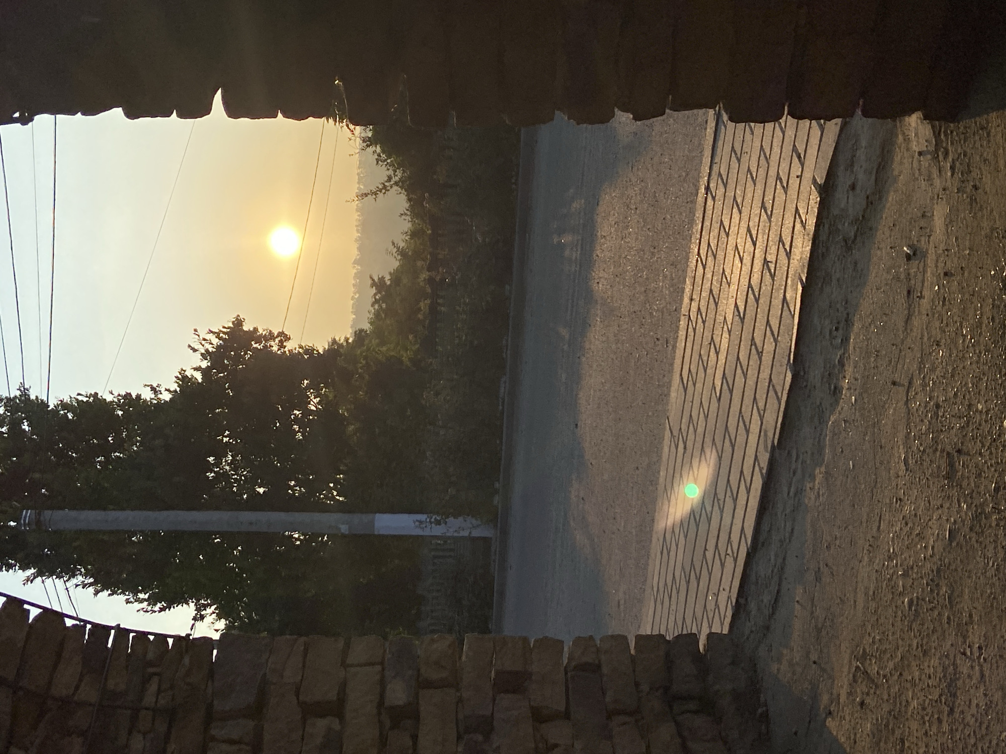 The image captures a street scene at sunrise, viewed through an arched stone structure. The sun, a bright orb, is low in the sky, casting a warm glow and long shadows across the pavement. The road and a brick-paved walkway are visible. On the left, a weathered stone wall forms the archway, with another stone pillar on the right. Beyond the road, a line of trees and foliage obscures the background, with a utility pole standing tall amidst the greenery. The sky is a pale yellow, indicating the early morning light. There are no people or vehicles present in the scene.