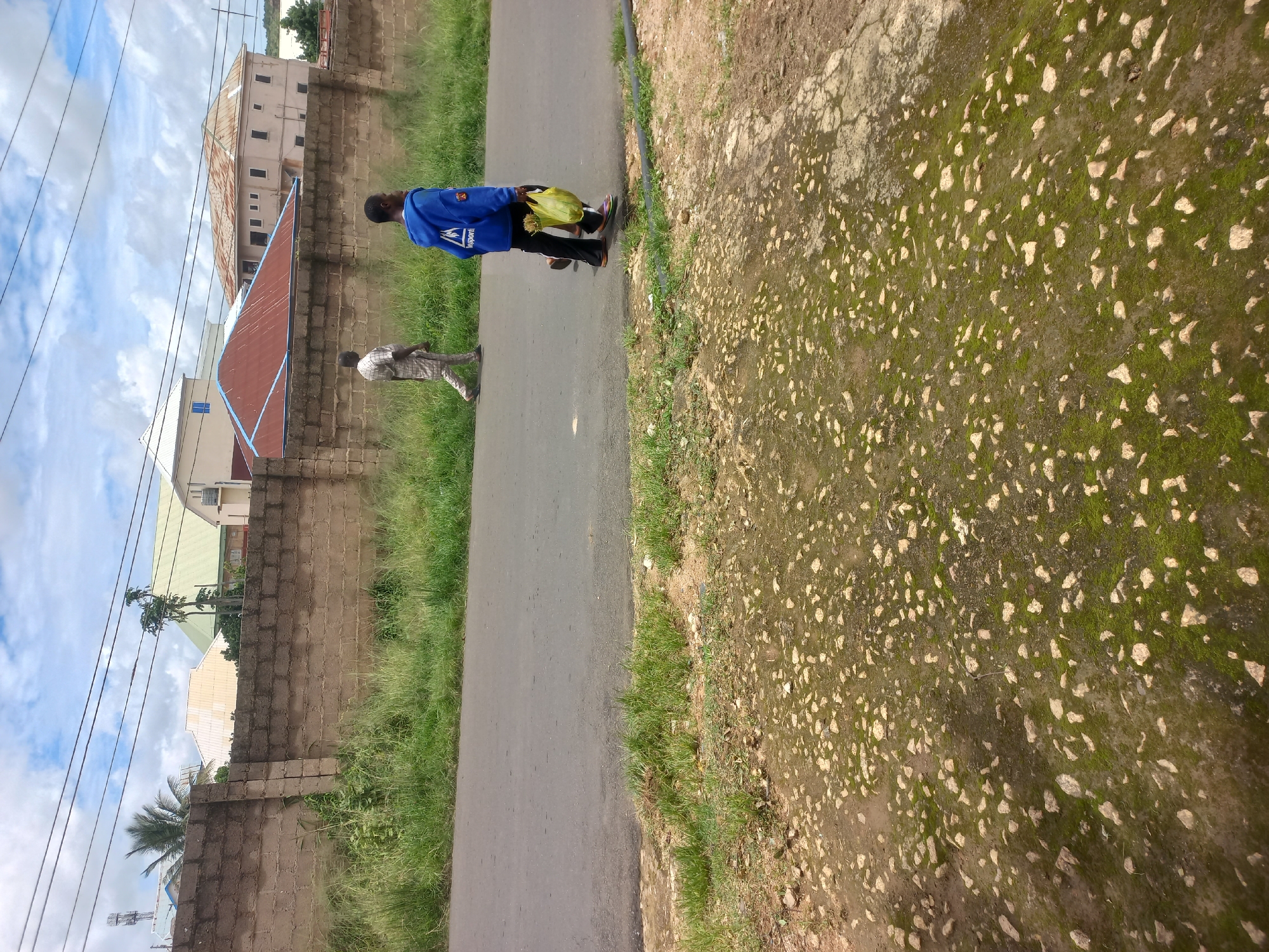 Two men walk on a paved road. The man on the left wears a striped tunic and pants. The man on the right wears a blue hooded sweatshirt and black pants, carrying a yellow plastic bag containing produce in his right hand. They are walking away from the camera. Behind them is a concrete block wall, overgrown with grass. Beyond the wall are several buildings with corrugated metal roofs, some painted red and others rust-colored, and a multi-story building with a light-colored facade. Palm trees and other vegetation are visible behind the wall. The ground in the foreground is unpaved and covered with rocks and patches of moss. The sky is cloudy.