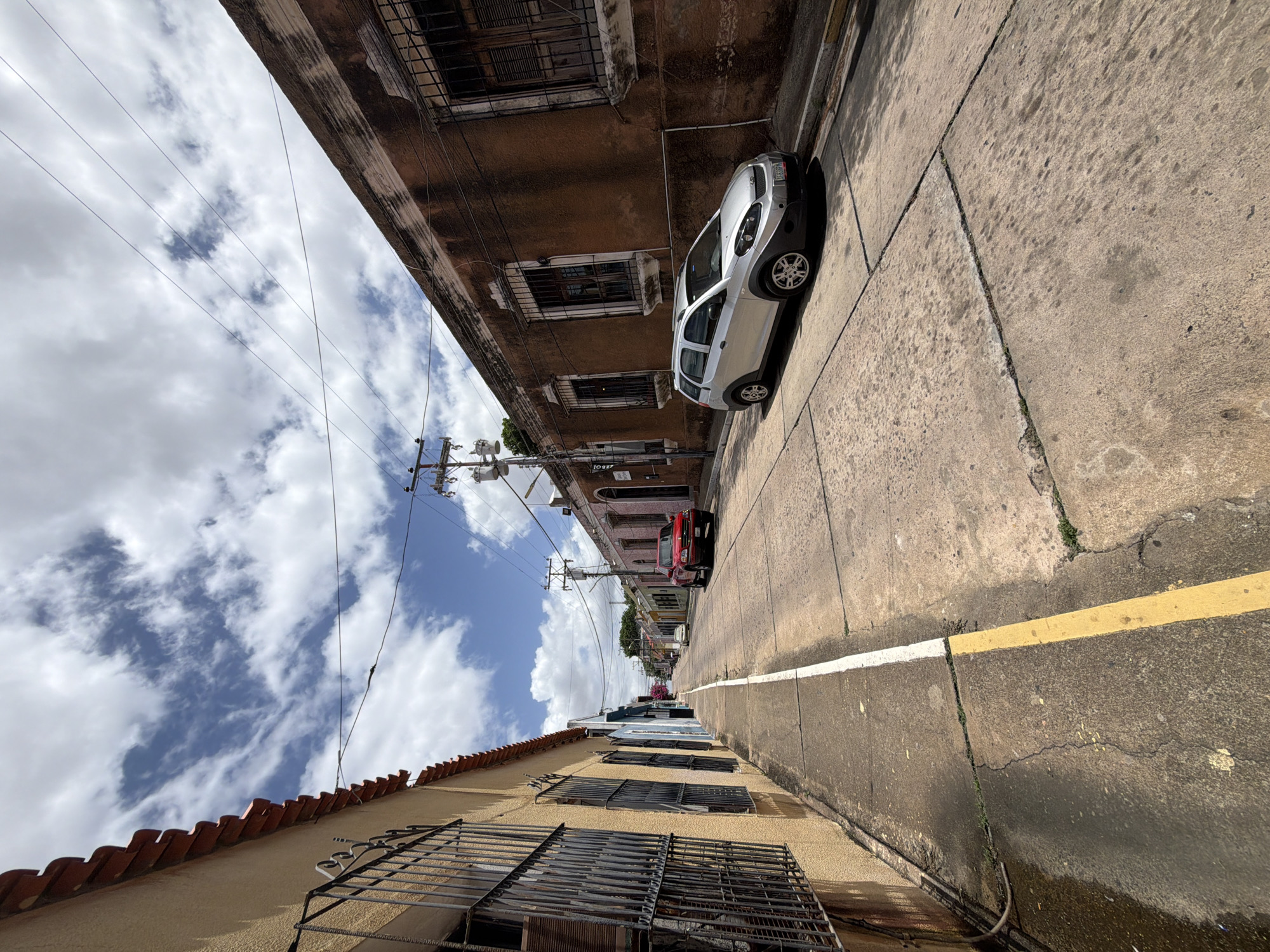 The image depicts a street scene in Ciudad Bolivar, Venezuela. A silver compact car is parked on the right side of the street, close to a brown building with barred windows. Further down the street, a red car is also parked. The street has a white line marking the edge, with a yellow line next to it. The sky is visible with scattered clouds and power lines stretch overhead. The buildings on either side of the street show signs of age and varying architectural styles, including a tan building with a red-tiled roof and barred windows.