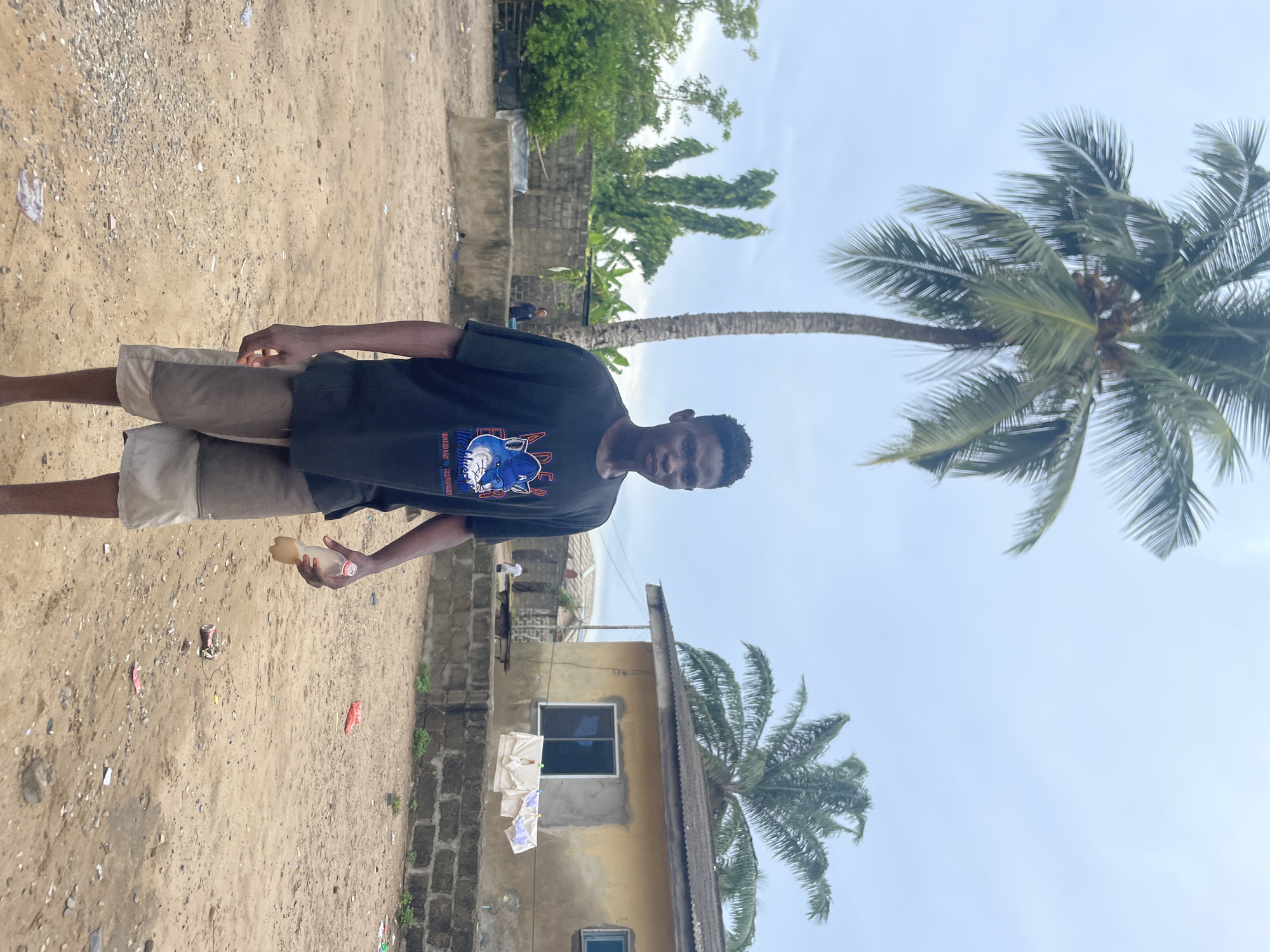 This image captures a young man standing outdoors on a sandy or dirt ground under a bright, overcast sky. The setting appears to be a residential or semi-rural area, consistent with Badagry, Nigeria.
The main subject is a young man with short, dark hair, positioned centrally and looking directly at the camera with a neutral expression. He is wearing a dark, possibly black, t-shirt featuring a blue and white graphic design on the chest. Visible text on the shirt includes "TREASURE," "THE WILD ONES," and partially readable words like "MAKING" and "MASTERS." He is also wearing light-colored, knee-length shorts, possibly beige and grey. In his right hand, he holds a clear plastic bottle with a red cap, which contains a yellowish liquid.
The ground beneath him is scattered with small pebbles and various pieces of litter, including plastic bags and wrappers. In the background, to the man's right, a low concrete block wall is visible. Beyond that, a simple, light-yellow painted building with a dark window frame and clothes hanging on a line can be seen. A prominent palm tree with a tall, slightly leaning trunk and lush fronds stands out against the sky. Additional green foliage and distant trees suggest a vibrant, natural environment. The time of day appears to be daytime, with soft, diffused light.