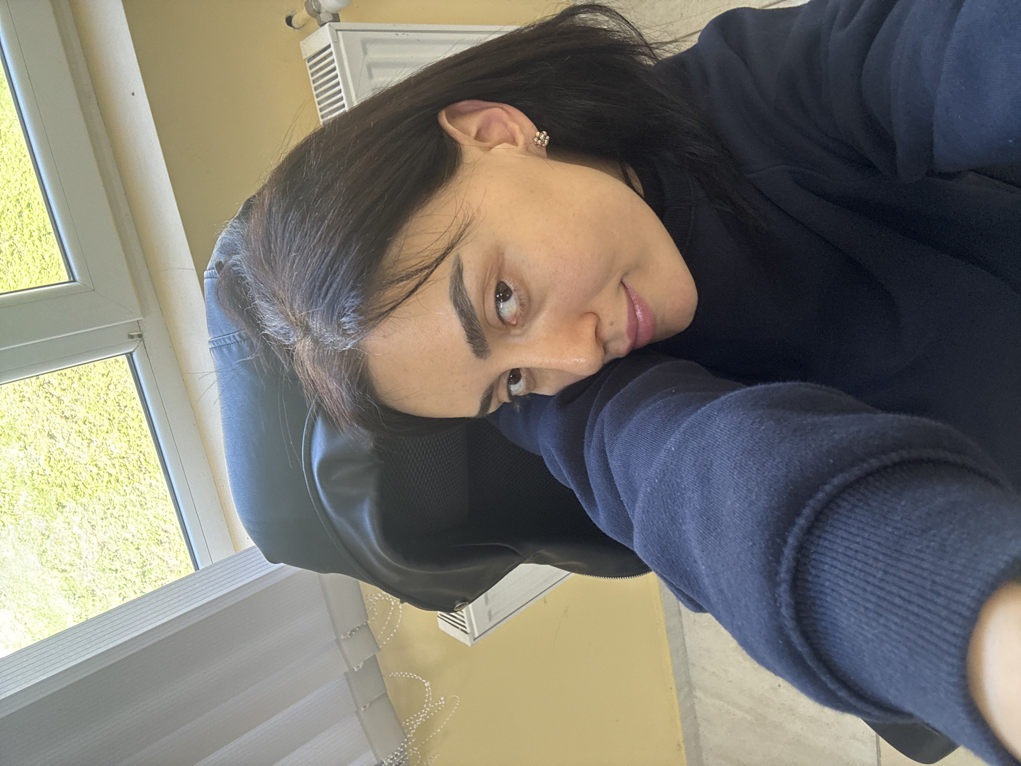 A woman with dark hair and brown eyes is leaning on a black leather jacket, resting against a black office chair. She wears a dark blue long-sleeved shirt and a small earring. The background features a window with white blinds and a view of greenery outside. A white air conditioning unit is visible on the wall. The scene appears to be indoors, and the overall lighting is soft. There is no indication of the location as Tbilisi, Georgia.