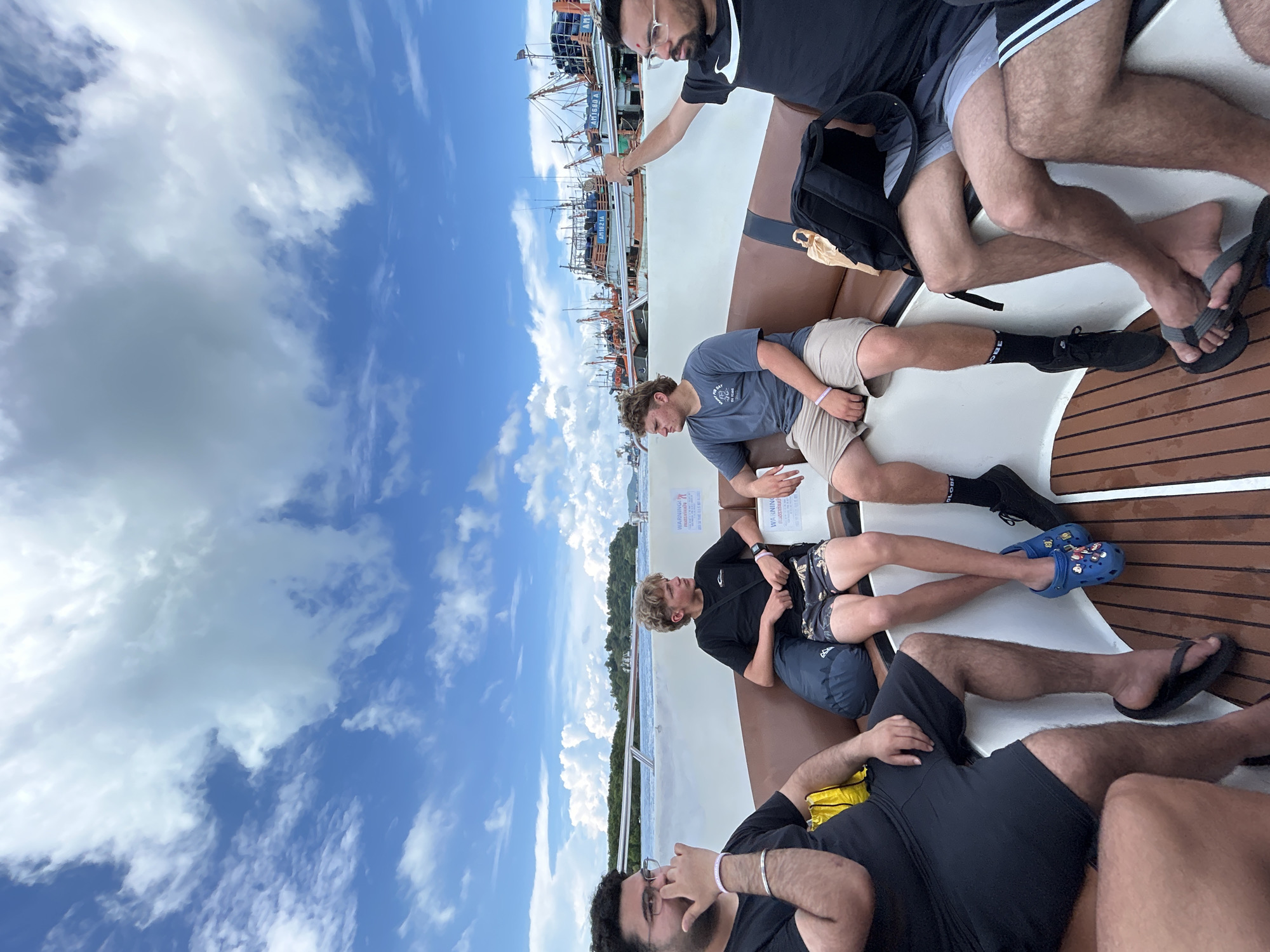 Four young men are seated on a boat in the ocean on a cloudy day, with a fleet of fishing boats docked in the background. The man on the far left is wearing glasses and a black t-shirt, with his hand covering his mouth. Next to him, a blonde-haired young man in a black t-shirt and patterned shorts is looking to the left. In the center, another young man in a grey t-shirt and tan shorts is looking forward. The man on the far right is wearing a black t-shirt and dark shorts, with his arm raised. The boat's interior is visible, featuring brown seating and a wooden floor. The scene appears to be in the waters near Ratsada, Thailand, given the presence of numerous fishing vessels common in the region.