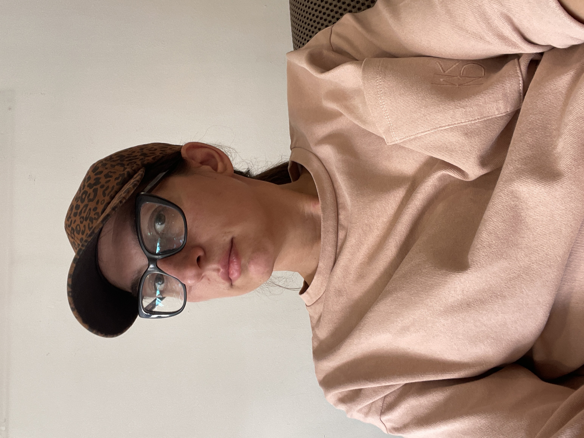 A close-up, waist-up portrait shows a woman wearing a pink long-sleeved shirt, a leopard-print baseball cap, and black-rimmed glasses. She is seated, and the background is a plain white wall. Her expression is neutral, and she is looking directly at the camera. The image was taken in an indoor setting, likely an apartment, in the city of Sofia, Bulgaria.