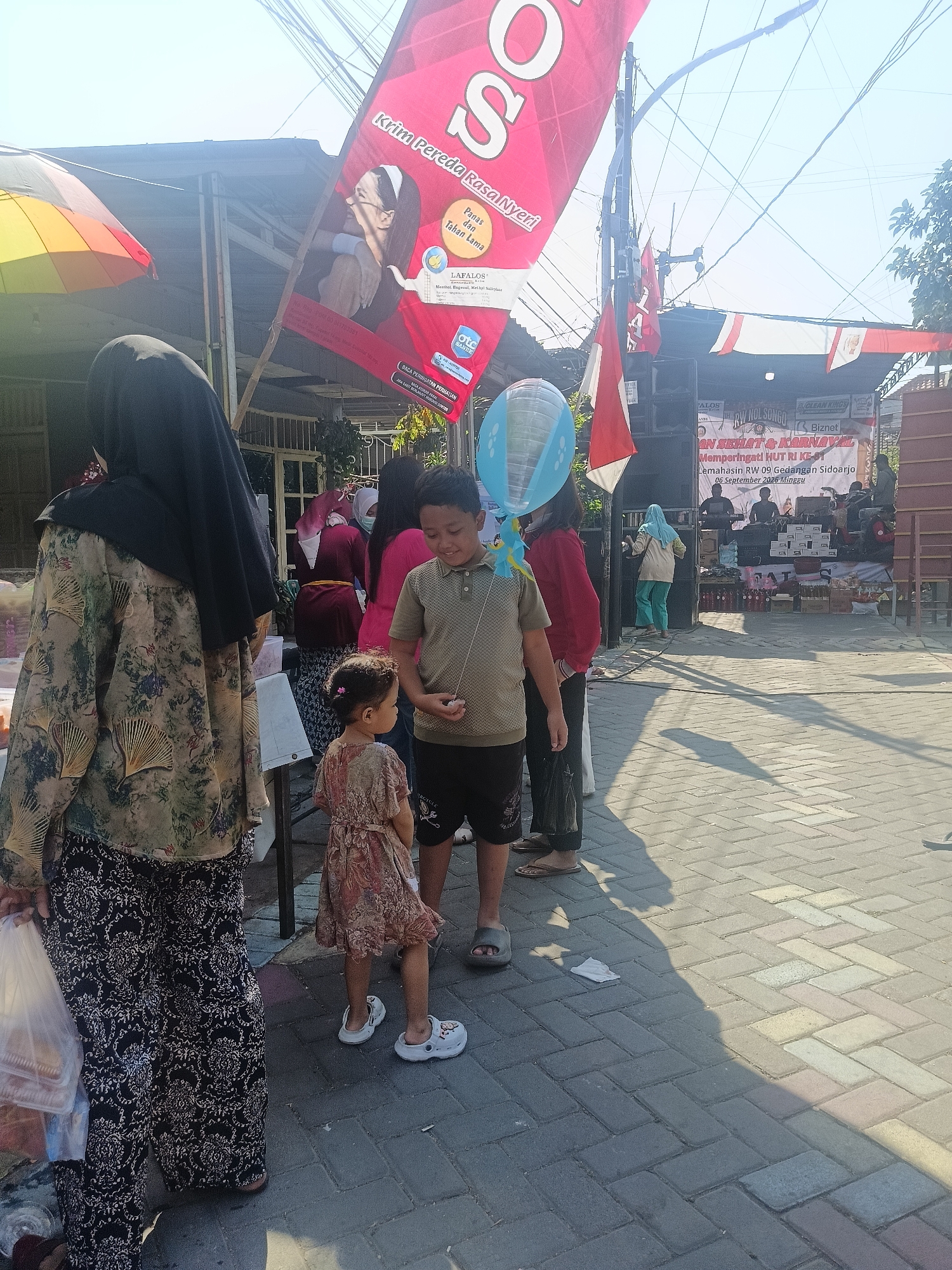 In Gedangan, Indonesia, an outdoor event is taking place on a brick-paved area, possibly a street or public square, under a bright, clear sky. A large red banner with white lettering, partially obscured, hangs from a pole, advertising what appears to be a product or event. A smaller red and white flag is also visible, along with power lines crisscrossing overhead.
In the foreground, a woman wearing a black hijab and a patterned dress walks away from the camera, holding a plastic bag. To her right, a young boy in a light brown t-shirt and dark shorts is holding a string attached to a blue balloon. He is looking towards the right of the frame. A young girl in a floral print dress and white clogs stands between the boy and the woman, facing right and looking slightly down. Her back is mostly to the camera.
In the middle ground, several other people are present, some in the background near a stage setup with speakers and equipment. A woman in a light blue headscarf and green pants is walking on the stage area. The environment suggests a festive or community gathering, possibly a street fair or local celebration given the banner, stage, and crowds. The sunlight casts long shadows on the brick pavement.