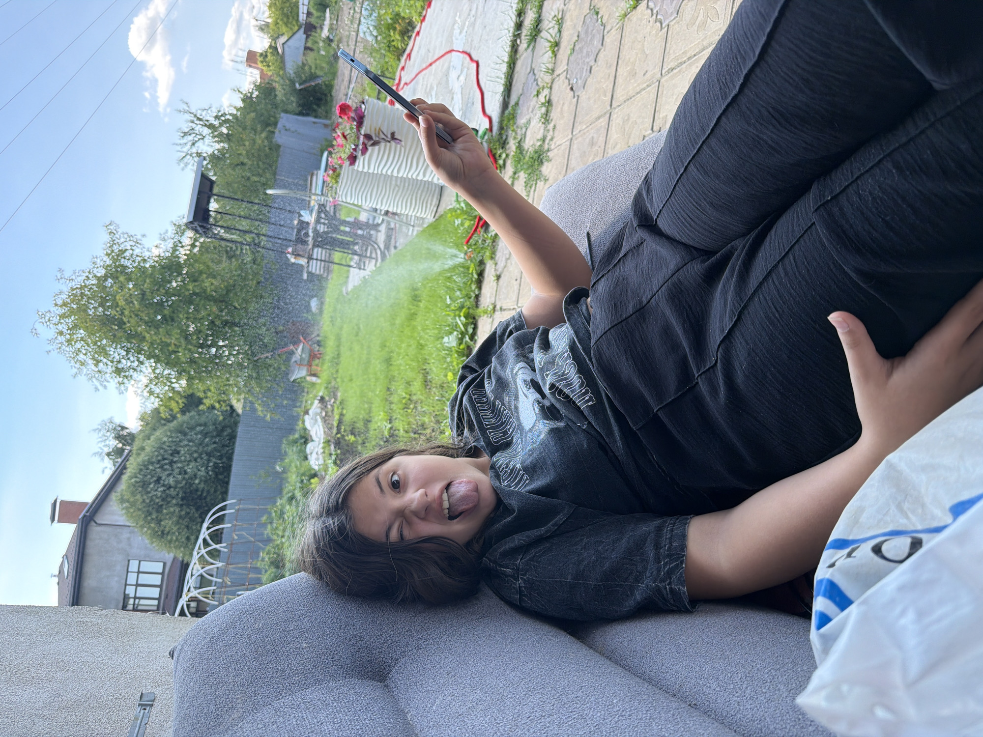 A young person with dark hair, wearing a dark t-shirt and dark pants, is lying on a gray sofa outdoors. The person is winking and sticking out their tongue while holding a smartphone. The background shows a yard with green grass, trees, a fence, and a house. There is a sprinkler system visible, watering the grass. The location appears to be a residential area in Gribki, Russia, likely a backyard given the presence of the house and garden elements.