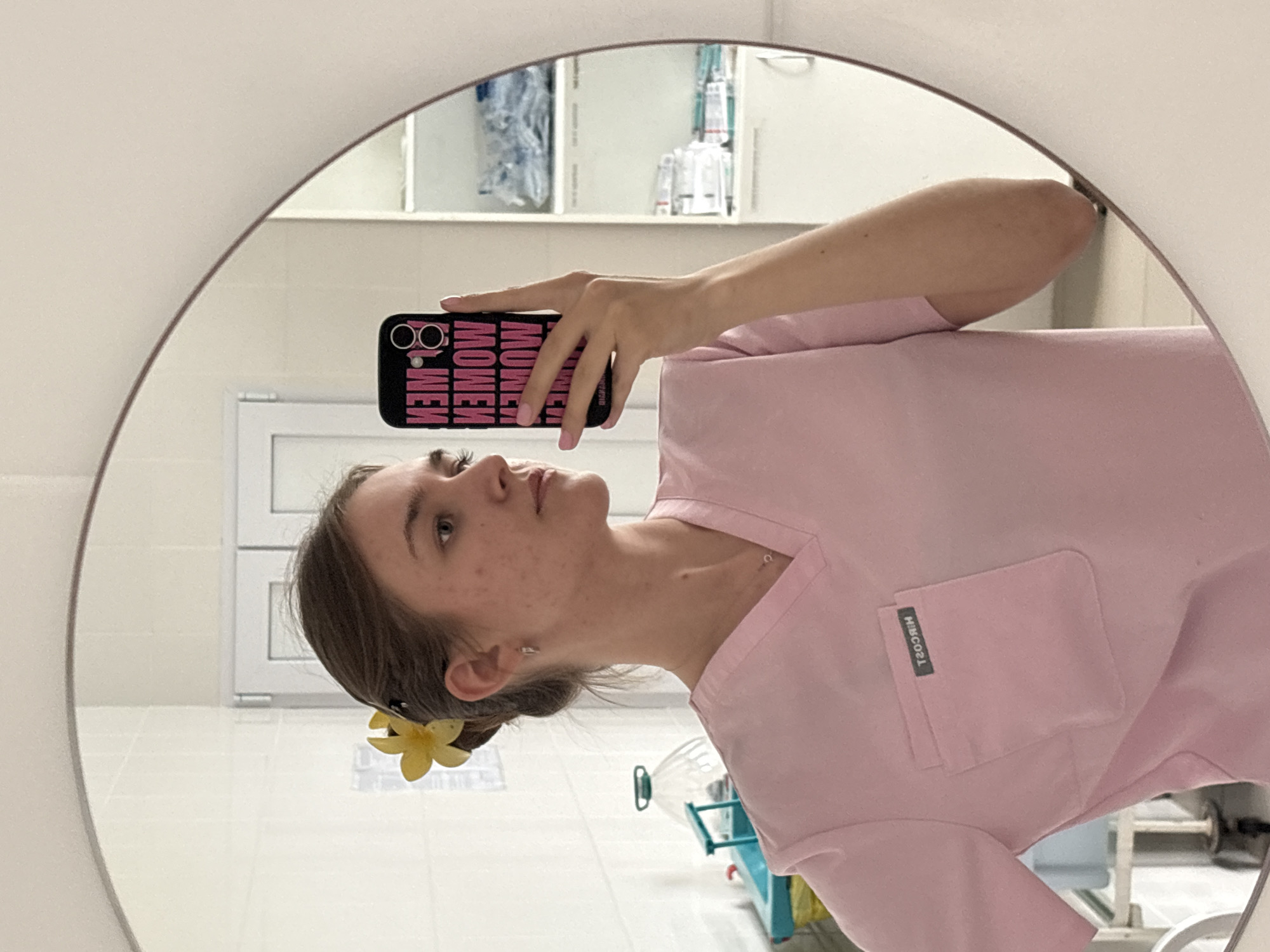 A woman is taking a mirror selfie. She is wearing a pink scrub top with a pocket on the right side. A yellow flower is clipped in her hair. She is holding a black cell phone with pink lettering. The reflection shows a bathroom setting, with a white door and cabinets in the background. The image likely originates from a medical or clinical setting, as suggested by the scrubs and bathroom environment. The location cannot be determined from the image content.