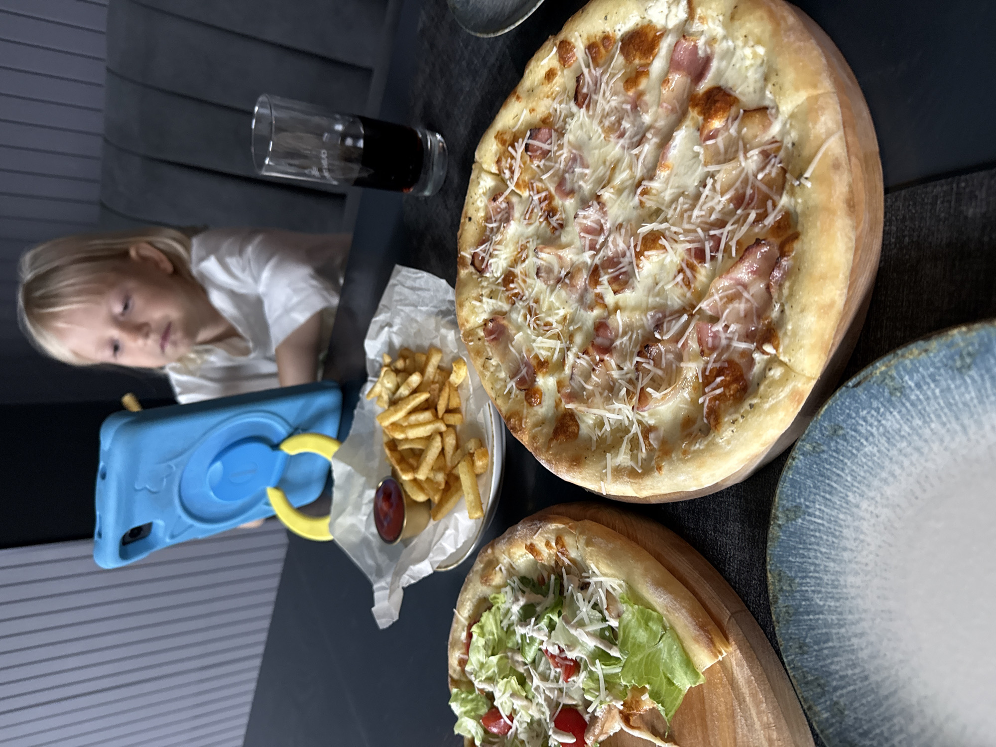 In Балаклавский округ, Ukraine, a young child with blonde hair sits at a dark table, engrossed in a blue tablet device. In front of the child, a glass of dark liquid, likely soda, stands beside a small plate of french fries with a dollop of ketchup. Two pizzas are prominently displayed on wooden boards: one topped with lettuce, tomatoes, and cheese, and another with bacon, cheese, and what appears to be a creamy sauce. A blue and white patterned plate occupies the foreground. The background features a dark, ribbed wall and a dark gray upholstered bench.