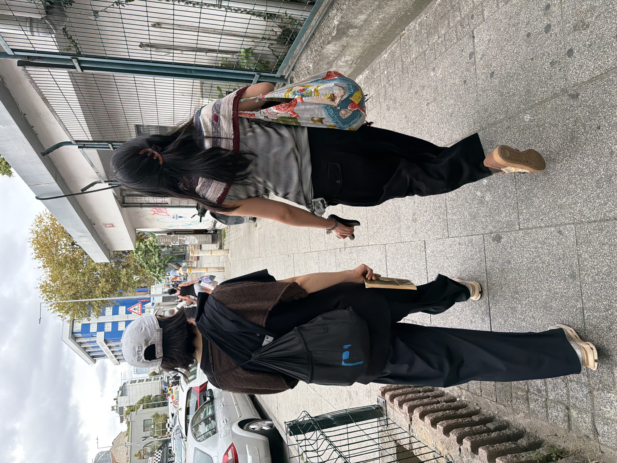 Two women walk away from the camera on a grey, paved sidewalk in Kadıköy, Turkey. The woman on the left wears a light grey baseball cap with the word "FOOTJOY" on it, a brown shawl over a black t-shirt, and loose-fitting black pants. She carries a black drawstring backpack with a blue graphic on it and holds a yellow book in her right hand. The woman on the right wears a striped sleeveless top with a patterned scarf draped over her shoulders and black pants. She carries a colorful patterned tote bag over her left shoulder. A metal fence with a chain-link pattern runs along the right side of the sidewalk, behind which is a white building with some graffiti. To the left of the sidewalk, there is a low brick wall and then a street with parked cars. The sky is overcast.