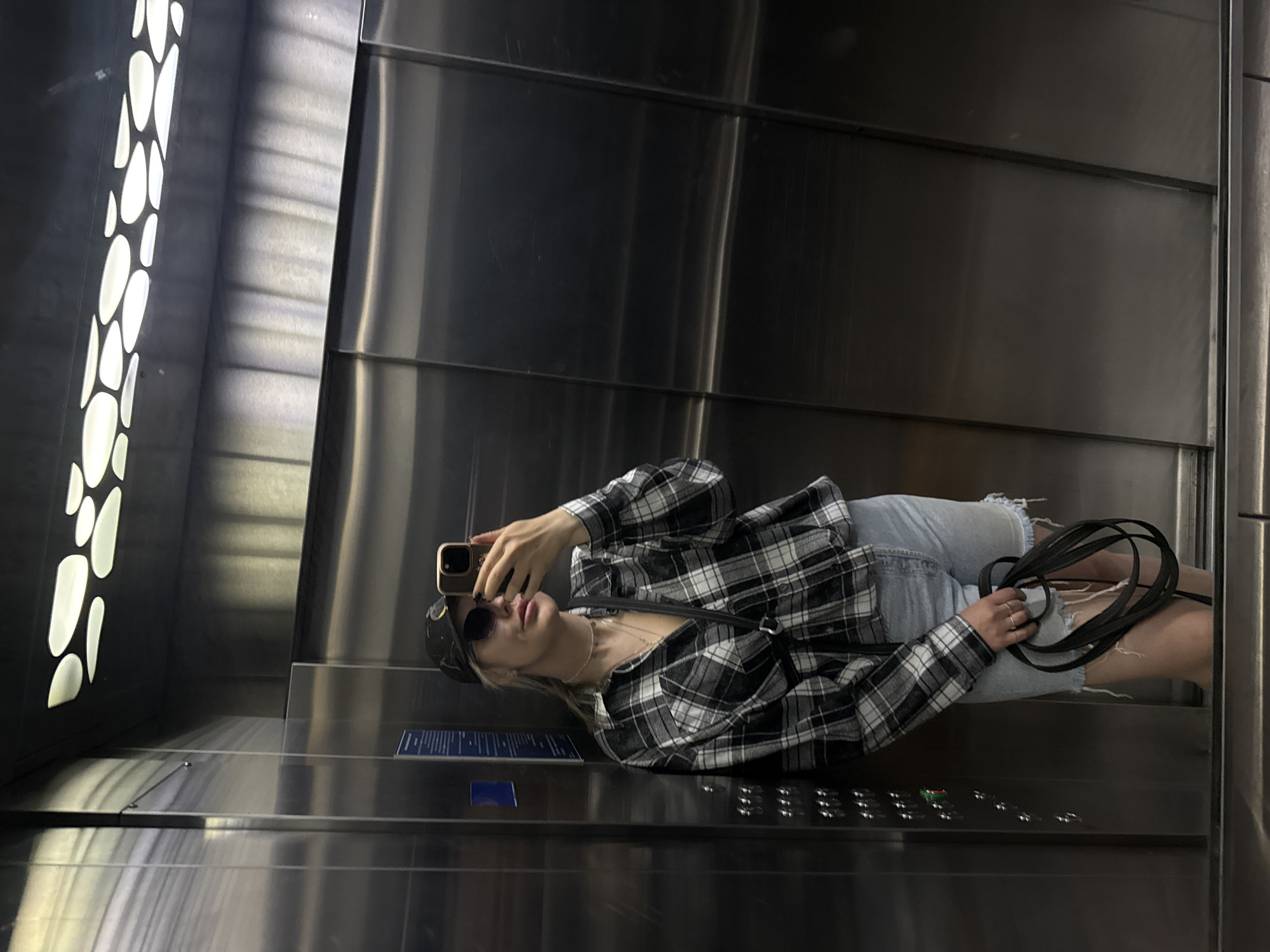 A person is captured taking a selfie in a modern, metallic elevator, in what appears to be Krasnoyarsk, Russia. The individual, seemingly female, is dressed in a black and white plaid button-up shirt, light blue distressed denim shorts, a dark baseball cap, and round sunglasses. They are holding a smartphone with a light-colored case up to photograph their reflection in the highly polished, metallic walls of the elevator. In their left hand, they hold a looped black strap, possibly from a bag or accessory.
The elevator interior features shiny, paneled metallic walls and a reflective floor. A prominent design element is a vertical light panel on the left side, featuring illuminated, irregular oval shapes against a dark background, casting a soft glow. A control panel with multiple silver buttons is visible on the bottom right, with one button displaying a green light. Above the control panel, a small blue plaque with illegible text is affixed to the wall. The overall lighting suggests an indoor, artificial environment.