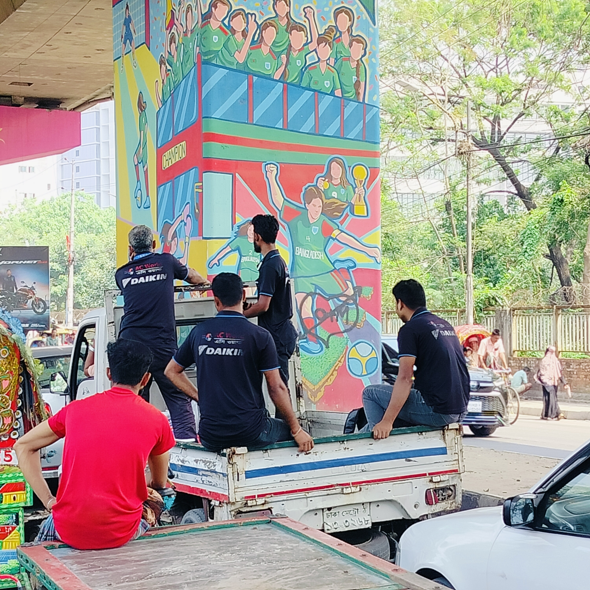 The image captures a street scene in Dhaka, Bangladesh. A small truck occupies the center of the frame, carrying five men. Four of the men wear navy blue t-shirts with the "DAIKIN" logo, indicating they might be employees of an air conditioning company. One of the men stands on the truck bed, appearing to be working on something above, while the other three are seated. A man in a bright red shirt sits on a decorative cart in the foreground.
Behind the truck stands a large concrete pillar adorned with a colorful mural. The mural depicts female athletes in green uniforms, celebrating a victory. The word "CHAMPION" is visible on the pillar. The athletes are identified as representing "BANGLADESH."
The background reveals typical urban features: a street with vehicular traffic, including a car and a rickshaw, and pedestrians walking by. Buildings and trees are visible in the distance. A motorcycle advertisement on a billboard can be seen on the left side of the image. The license plate of the truck reads "ঢাকা মেট্রো-ঠ ১৩-৩২৬৮" which is a Dhaka metro registration number.