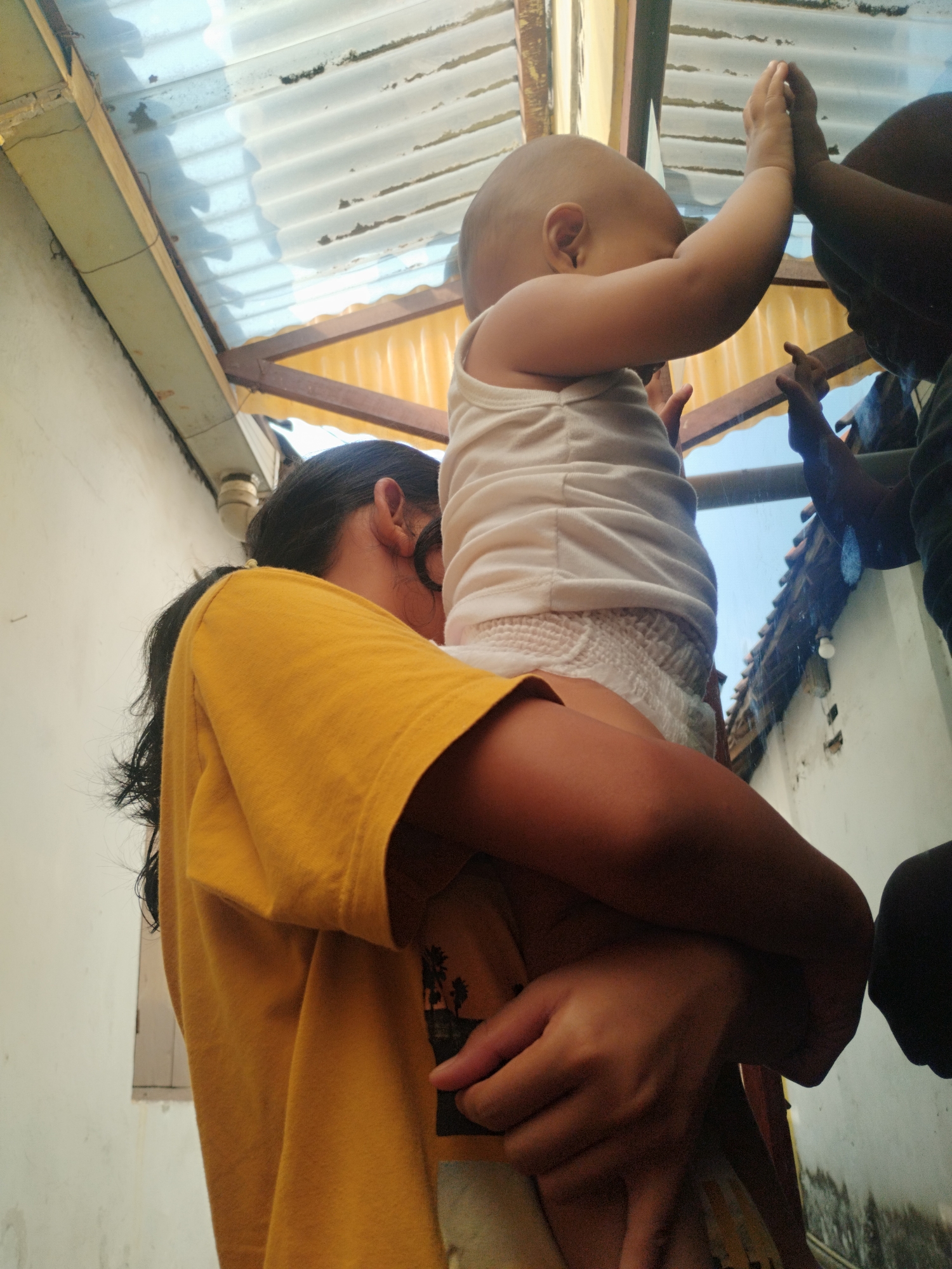 In Surabaya, Indonesia, a person is holding a baby in their arms, facing away from the camera. The baby is wearing a white tank top and a diaper, and its arms are raised. The person holding the baby is wearing a yellow t-shirt. They are standing under a corrugated metal roof, which is supported by wooden beams. The background shows a white wall and some greenery. The overall scene appears to be outdoors, possibly in a residential area, with a bright sky visible through the roof.