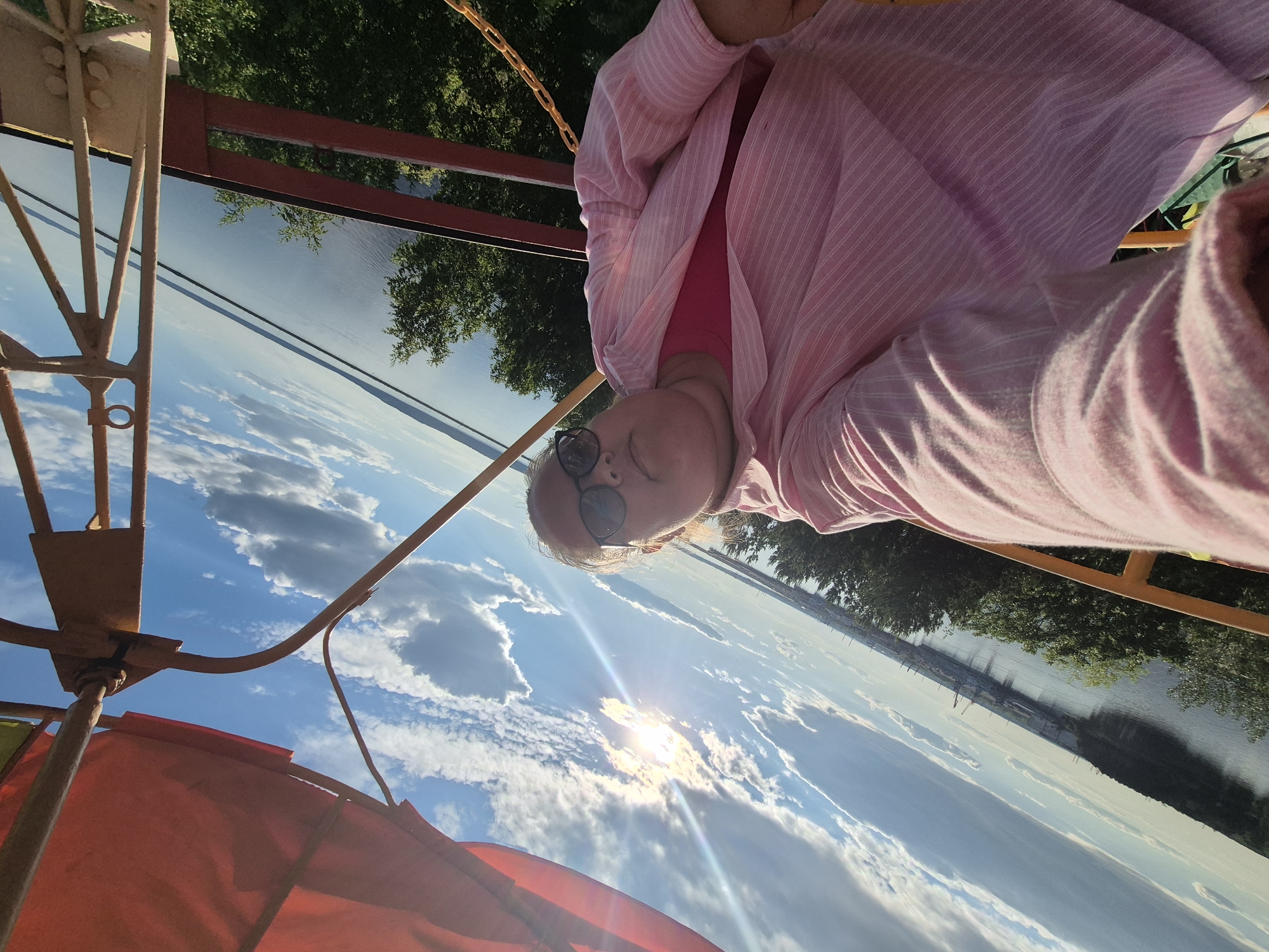 The image captures an adult female taking a selfie from an elevated vantage point, likely within a gondola or observation deck, identified by the surrounding rust-colored metal framework and a red fabric canopy visible in the upper left. The subject is depicted with fair skin, light-colored hair, and is wearing black-rimmed sunglasses. She is dressed in a light pink, vertically striped, long-sleeved button-up shirt worn open over a solid pink t-shirt. Her right arm is extended, holding the camera, with the hand partially visible in the lower right corner.
The background reveals an expansive body of water, characteristic of the Kama River, extending horizontally across the frame. The sky is clear blue with scattered white cumulus clouds, and the sun is positioned prominently in the upper left quadrant, creating a distinct lens flare. Along the distant left bank of the river, an industrial port is visible, characterized by numerous cranes and structures. The right bank is densely covered with green trees. The image has a significant diagonal tilt, suggesting an angled shot or a rotating structure. The scene is consistent with views over the Kama River in Chaikovsky, Russia, potentially from a Ferris wheel or an observation platform overlooking the Chaikovsky Hydroelectric Power Station and its surroundings.