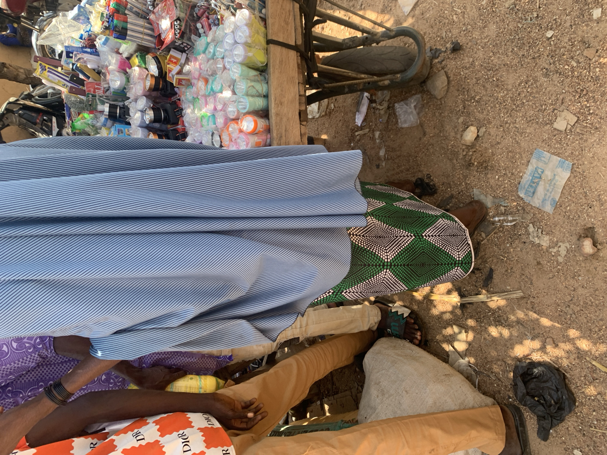 The image depicts an outdoor market scene in Boi A, Nigeria. The foreground is dominated by the legs and lower torsos of several people, suggesting a close-up perspective of individuals engaged in or observing the market activity. One person is wearing a light blue and white striped garment, partially covering their lower body. Another is dressed in a purple patterned fabric. A third person is wearing tan pants. The ground is sandy and littered with debris, including plastic bags and some dry vegetation.
In the background, a cart is laden with an assortment of items, including what appear to be flashlights or small lamps packaged in plastic, pens, and various other goods. A bicycle wheel and part of its frame are also visible, suggesting that the cart might be attached to or near a bicycle. The lighting and shadows indicate it is daytime, likely during the morning or afternoon. The overall impression is one of a bustling, informal marketplace, characteristic of many communities in Nigeria. The arrangement of goods on the cart implies that a sale or trade is in progress, or about to commence.