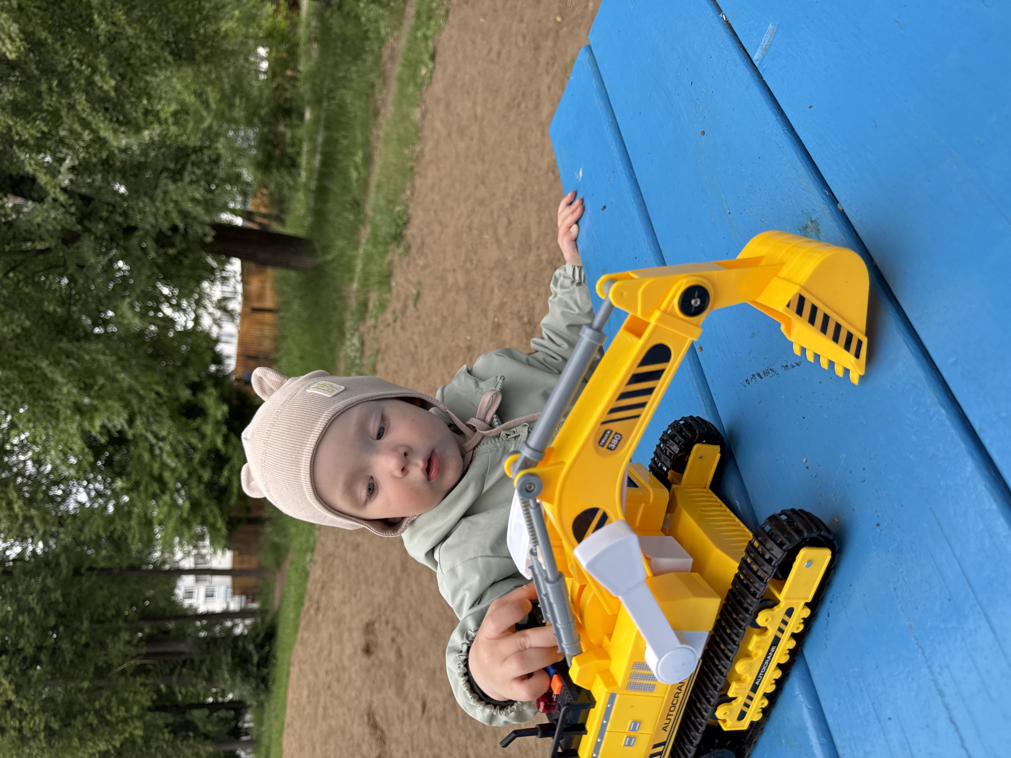 A young child wearing a beige hat with bear ears sits at a blue picnic table and plays with a yellow toy excavator. The child's left hand is on the toy's controls, and their right hand rests on the table. The excavator is positioned on the left side of the table, with its boom extended. The background shows a sandy playground area with trees and a building. The setting appears to be an outdoor playground in Chaikovsky, Russia.