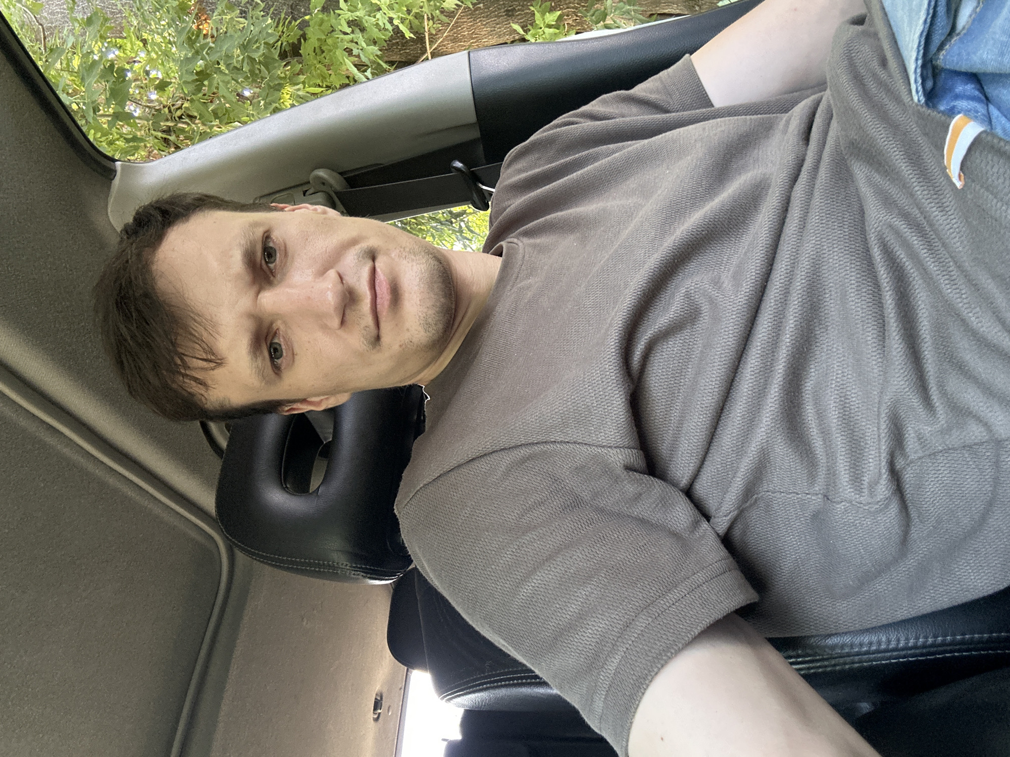 A man sits in the driver's seat of a vehicle, with his head and upper body filling the frame. He is wearing a short-sleeved, taupe-colored t-shirt and denim jeans. The interior of the car is visible, including the headrest of the seat behind him, the sunroof above, and the car's interior trim. Outside the car, through the window, green foliage and trees are visible. The lighting suggests it is daytime, possibly late afternoon. The image appears to have been taken by the man himself, indicated by the angle and framing. The location is identified as Makiv, Ukraine.