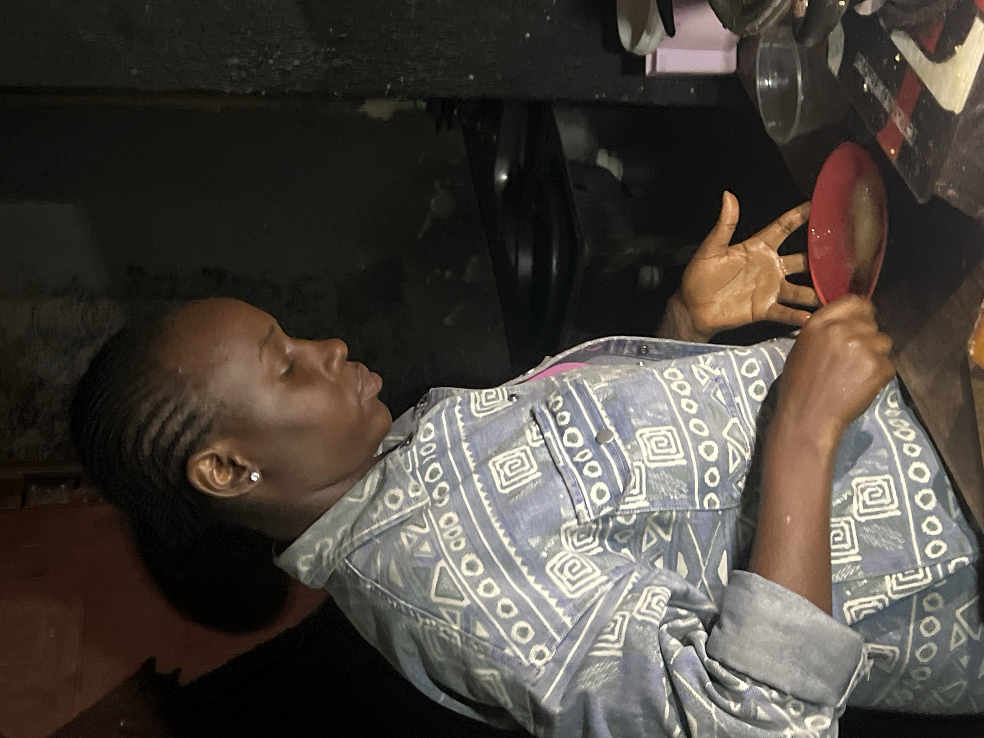 A young Black woman with braided hair is depicted in a dimly lit indoor setting, likely a kitchen or food preparation area. She is wearing a blue and white patterned jacket over a pink garment. Her head is bowed as she looks down at a small red bowl in her right hand, which she is holding over a dark surface. Her left hand is resting on the same surface. In the background, a dark stovetop is visible, along with some other kitchen items. The overall impression is of a moment of focused activity. The location is described as Durumi, Nigeria.