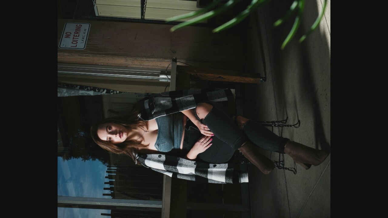 A young woman with long brown hair and fair skin is seated on a metal stool in front of a glass-paned window. She is wearing a denim bustier top, a black and white plaid shirt worn open, and dark, ripped jeans. Her legs are crossed, and she is wearing brown heeled boots. She is looking directly at the camera with a neutral expression. The background appears to be an urban setting, possibly a cafe or an industrial area, with a table and chairs visible behind her. A sign that reads "NO LOITERING" is affixed to a concrete pillar to her right. The lighting suggests it is dusk or evening, with shadows cast on the ground.