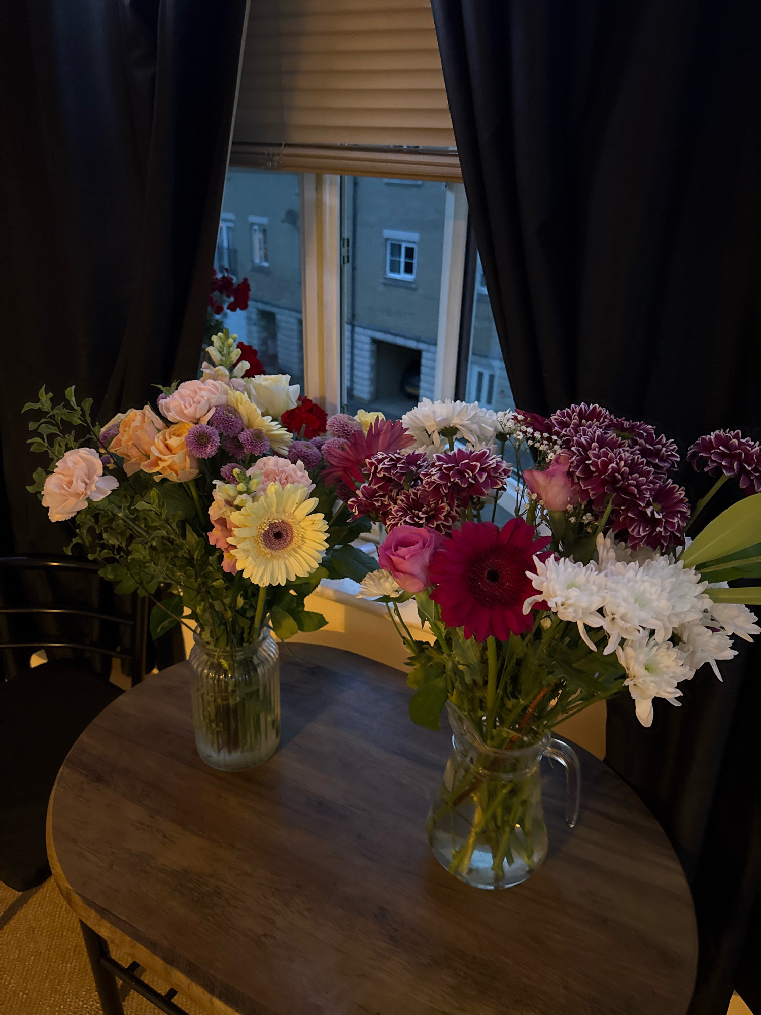 This is a medium-angle, eye-level, indoor photograph taken in the city of Great Yarmouth, United Kingdom. The scene depicts two glass vases filled with a variety of colorful flowers, placed on a round, wooden table. The table is situated in front of a window with a black curtain drawn to the left and right sides, partially obscuring the view. Through the window, buildings with windows and a balcony are visible, suggesting an urban environment. The lighting appears to be dim, with artificial light sources casting soft illumination on the flowers and table. No people are present in the frame. The flowers in the left vase include pink and peach roses, a yellow gerbera daisy, and purple filler flowers. The right vase contains white chrysanthemums, deep pink gerbera daisies, and dark purple chrysanthemums with white edges. The overall impression is one of a domestic setting, possibly a home or a quaint establishment in Great Yarmouth.