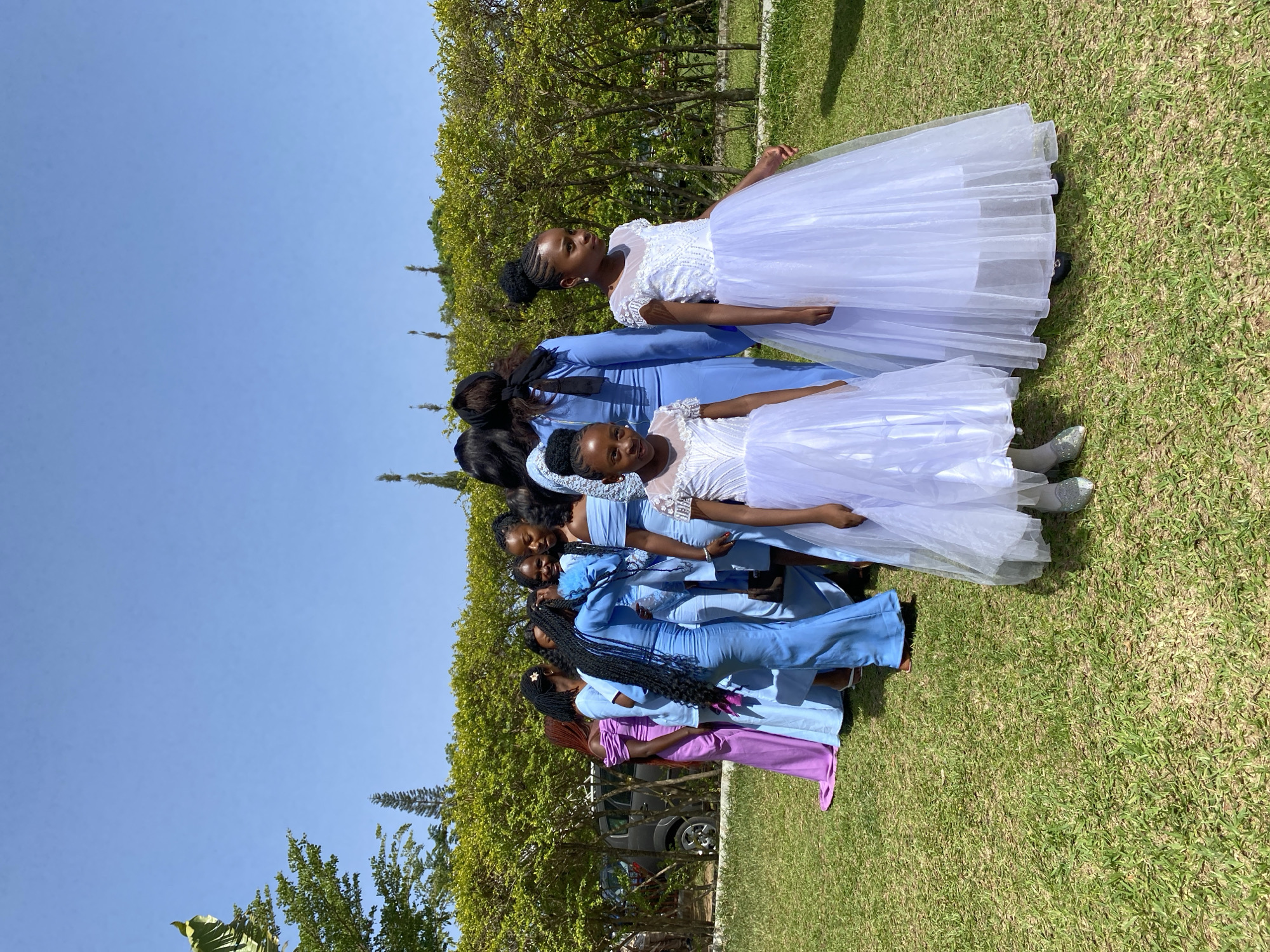 A group of approximately 10 young women and girls are positioned closely together on a grassy lawn under a clear blue sky. The location appears to be in a natural setting, evidenced by nearby trees and vegetation. The women and girls are dressed in a mix of formal attire: some wear long, flowing gowns in shades of light blue and purple, while others are in white dresses with tulle skirts. The individuals are arranged in a line, facing the same direction. In the background, a vehicle is partially visible. No explicit actions or interactions are immediately apparent; the group seems posed for a photo. Given the available information, it is not possible to verify the precise location of this photograph in Bukuru, Nigeria.