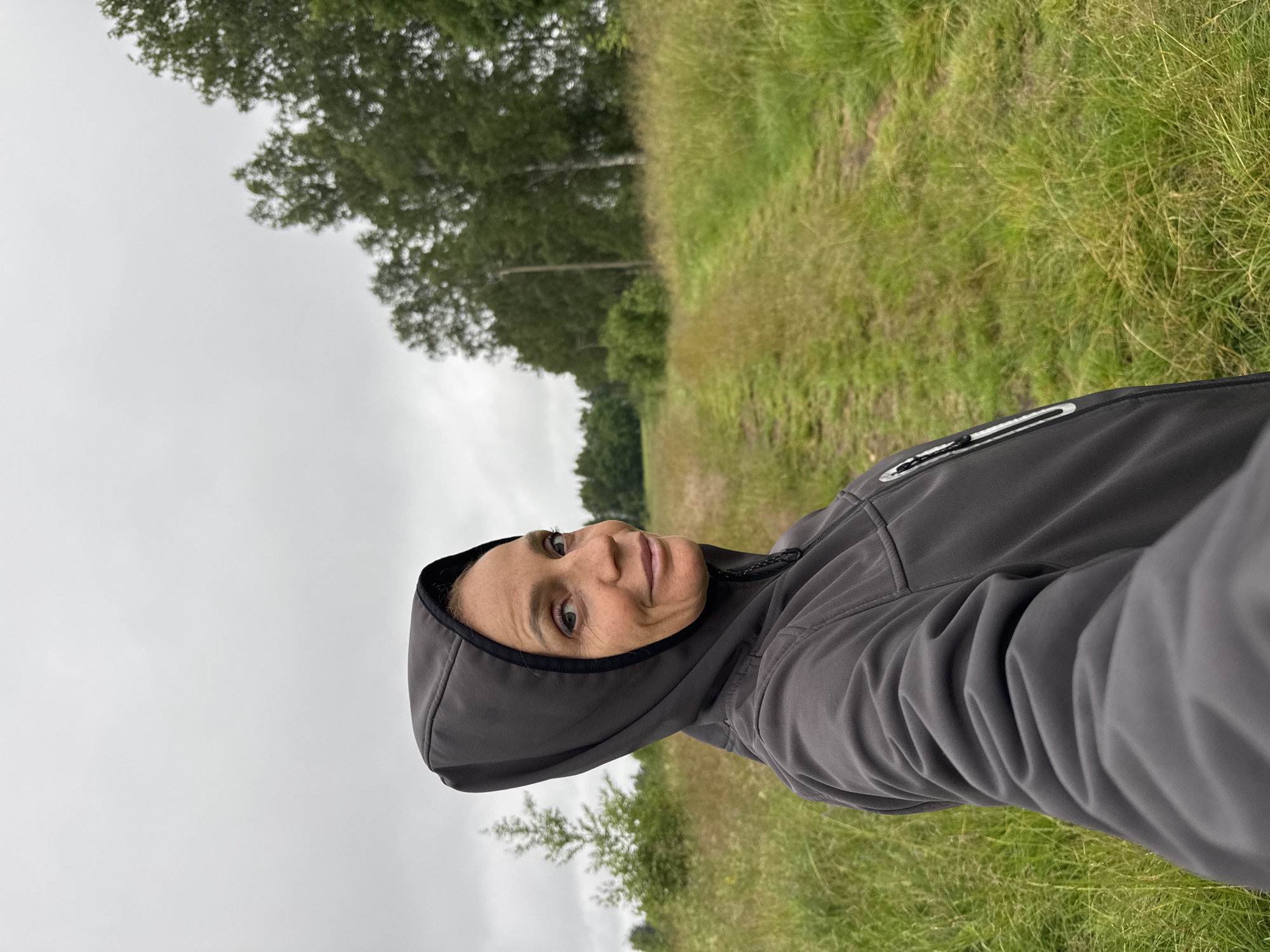 The image shows a woman in a gray hooded jacket, taken in a selfie format. She is outdoors, with a grassy field and trees in the background. The sky is overcast, suggesting a cloudy or rainy day. The woman is smiling and looking at the camera, her face is well-lit. The setting appears to be a rural or natural environment, possibly a park or a trail, given the presence of a path running through the grass. The orientation of the image is portrait, with the subject's head towards the top left of the frame and her body extending towards the bottom right. There is no visible text in the image. The location context provided is Mars, Russia.