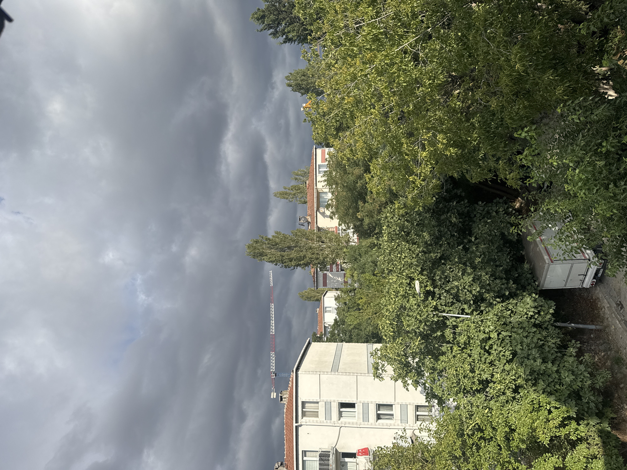 The image shows a view of apartment buildings and trees under a dark, cloudy sky, likely in the Gungoren district of Istanbul, Turkey. A construction crane is visible in the distance, stretching across the frame above the buildings. The foreground is dominated by lush green trees, with a white truck parked on a paved area below. One of the apartment buildings has a red and white flag displayed on its facade. The overall lighting suggests an overcast day, possibly before or after a rain shower.