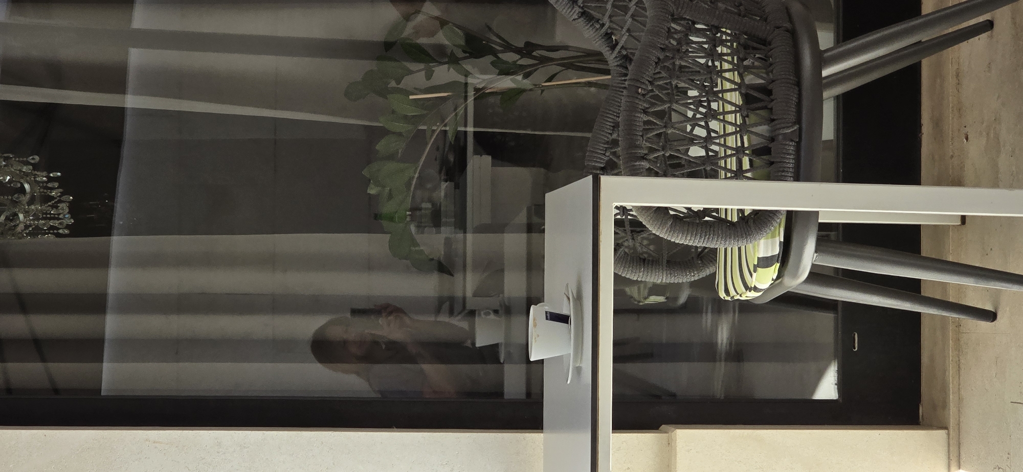 A reflective window, possibly in Budva, Montenegro, offers a view into a dimly lit interior and a partial reflection of the exterior. In the reflection, a person is visible, holding a smartphone and taking a picture. In the foreground, a white table holds a white coffee cup with a blue stripe on a saucer. To the right, a grey woven chair with a green and white striped cushion is visible. The background features sheer curtains and a potted plant with dark green leaves. The overall scene suggests a casual setting, perhaps a cafe or a home.
