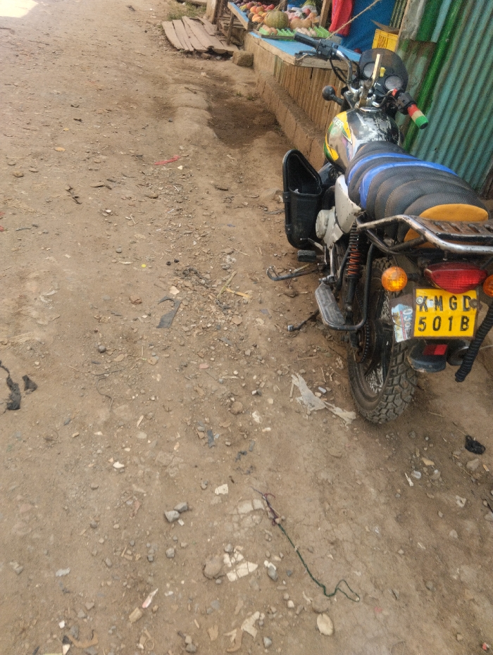 This image depicts a static outdoor scene within Witeithie ward, Kenya, characterized by an unpaved ground surface and commercial activity.
**Environment:** The foreground and middle ground consist of a dirt path, sparsely covered with small stones, gravel, and scattered debris including bits of plastic, paper, and a green-and-purple string-like object extending across the lower-middle section. A subtle depression or channel in the dirt runs parallel to the right edge of the frame, suggesting a drainage path.
**Objects:**
* **Motorcycle:** A black, yellow, and green motorcycle is parked on the dirt path on the right side of the frame, facing away from the viewer at a slight angle. It is equipped with a black seat featuring blue horizontal ribbing and a smaller yellow/orange section at the rear. A black plastic storage container is attached to its left side, open. The rear of the motorcycle displays a yellow Kenyan license plate with black lettering: "K M G D / 501 B", flanked by two round orange turn signal lights. The handlebars show details of green and red grip elements. The tires exhibit a knobby tread pattern.
* **Market Stalls:** Along the right side of the path, parallel to the motorcycle, a market stall is visible. It features a light blue counter on which various fruits and vegetables are displayed, including a large pumpkin-like gourd and other green and reddish produce items. Behind this counter is a vertical corrugated metal wall, colored greenish-blue. Further down the path, beyond the motorcycle, wooden planks are visible, possibly forming part of another stall or ground covering.
**People/Actions/Interactions:** No people are visible in the scene. The motorcycle is stationary, and no actions or interactions are depicted.