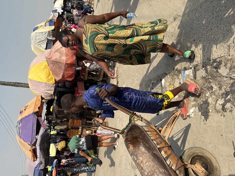 The scene depicts an outdoor market in Ado, Nigeria, under clear, bright daylight. In the foreground, a young male, appearing to be of African descent, walks left-to-right pushing a single-wheeled, rusted wheelbarrow. He wears a dark blue tunic over yellow patterned shorts and red slides with "CCI" branding, holding the wheelbarrow's green-wrapped handles. To his right and slightly behind, an adult female, also appearing to be of African descent, walks in the same direction, holding a clear plastic water bottle in her right hand. She wears a patterned yellow, green, and brown strapless dress and a single green slide on her left foot. The paved ground beneath them is marked by scattered debris, including plastic and other refuse. In the background, numerous market stalls are visible, sheltered by a canopy of large, multicolored umbrellas (yellow, orange, purple, pink, white). Goods, including stacked produce, are displayed at these stalls. Multiple other individuals are present throughout the market background, moving and interacting. Overhead power or communication lines are visible against the pale blue sky.