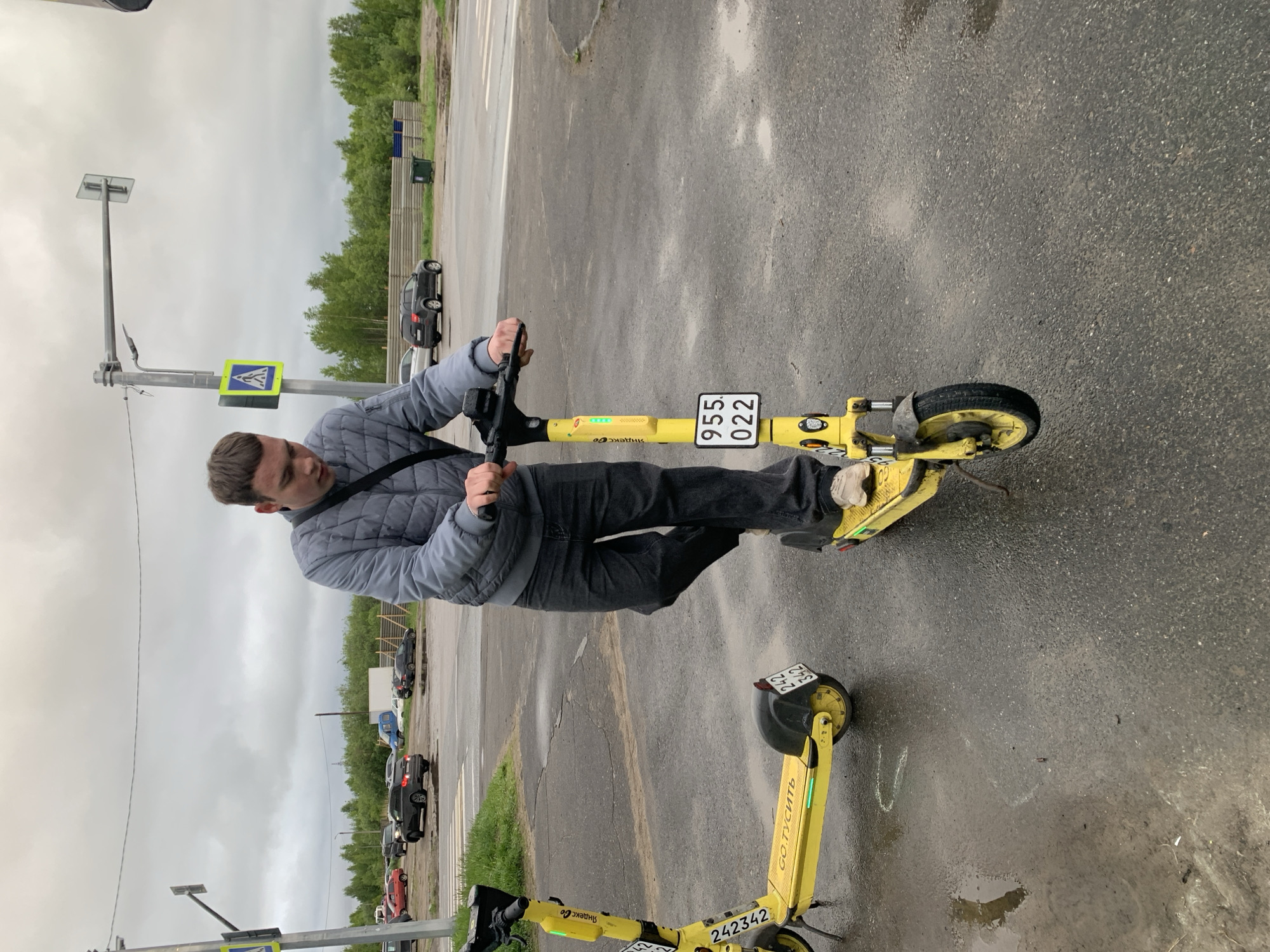 A man is depicted standing on a bright yellow electric scooter in an outdoor paved area, possibly a parking lot or roadside, in Severodvinsk, Russia. He is dressed in a grey quilted jacket, dark pants, and light-colored shoes, holding the handlebars as if preparing to ride or having just stopped.
Another identical yellow electric scooter is visible parked in the foreground. The ground is dark, wet asphalt with small puddles, indicating recent rain. The sky is overcast and grey, suggesting a cloudy daytime. In the background, a line of cars is visible, some parked and others possibly moving, with a treeline and a wooden fence further behind. A tall pole to the left features a pedestrian crossing sign (blue square with a white triangle and black pedestrian symbol).
The scooter the man is on has the identifier "955 022" on a plate at the front and "Яндекс Go" (Yandex Go) branding on its stem, with several green indicator lights illuminated. The other scooter also shows "242 342" on its front plate and "GO ТУСИТЬ" (GO PARTY/HANG OUT) written along its side.