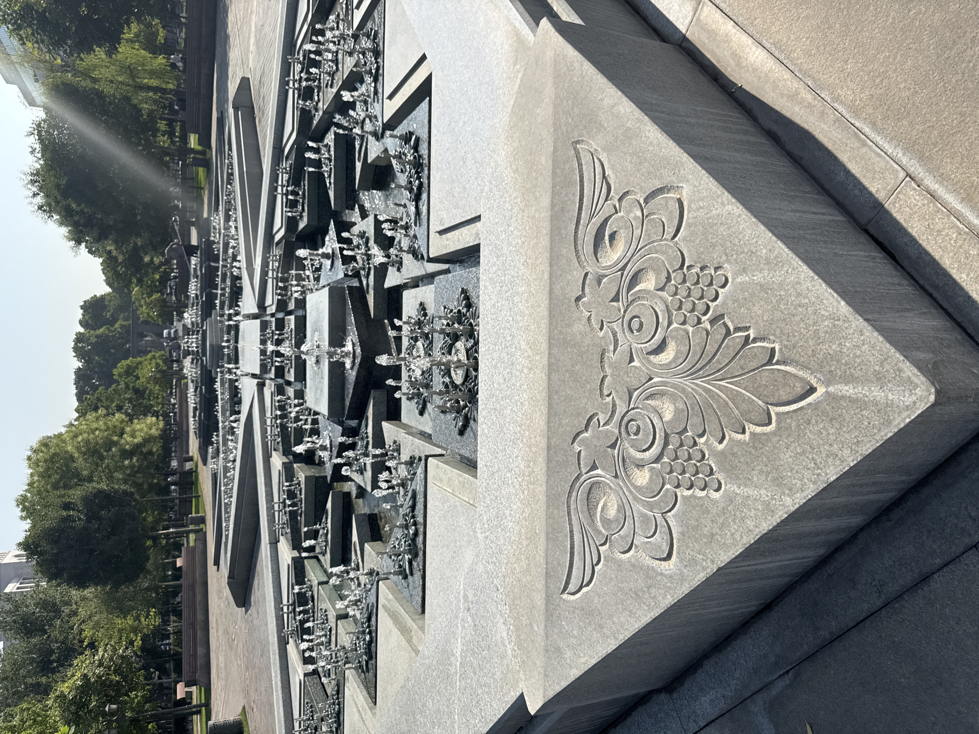 A wide-angle shot captures a large, multi-tiered fountain complex in Yerevan, Armenia, under a bright, sunny sky with a visible sunbeam effect. The foreground prominently displays a section of the fountain's granite base, intricately carved with a symmetrical floral and grape motif. The fountain itself is composed of numerous geometric concrete platforms and pools, from which multiple jets of water spray upwards. The water streams create a dynamic, shimmering effect against the starkness of the stone. In the background, a park setting unfolds with lush green trees lining paved pathways and benches, suggesting a public space. The overall impression is one of a meticulously designed urban landscape feature, showcasing both natural elements and architectural artistry.