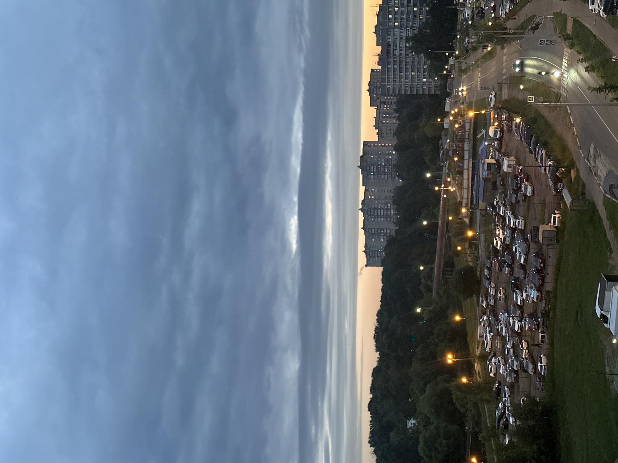 The image depicts an overcast twilight scene in Sverdlovskiy, Russia. In the foreground, a parking lot filled with numerous cars is visible, with streetlights casting a warm glow. Beyond the parking lot, a dense line of trees forms a dark green backdrop, with a red bridge arching over a dark area, possibly a river or ravine, in the middle distance. Further back, a cityscape with multi-story apartment buildings, illuminated with numerous windows, stretches across the horizon. The sky is dominated by heavy, layered gray clouds with streaks of pale orange and yellow light near the horizon, indicating the lingering twilight. A road with moving vehicles, including cars and a motorcycle, is visible on the right side of the image, bordered by sidewalks and more apartment buildings. The overall atmosphere is calm and urban, with the natural elements of trees and sky contrasting with the built environment.