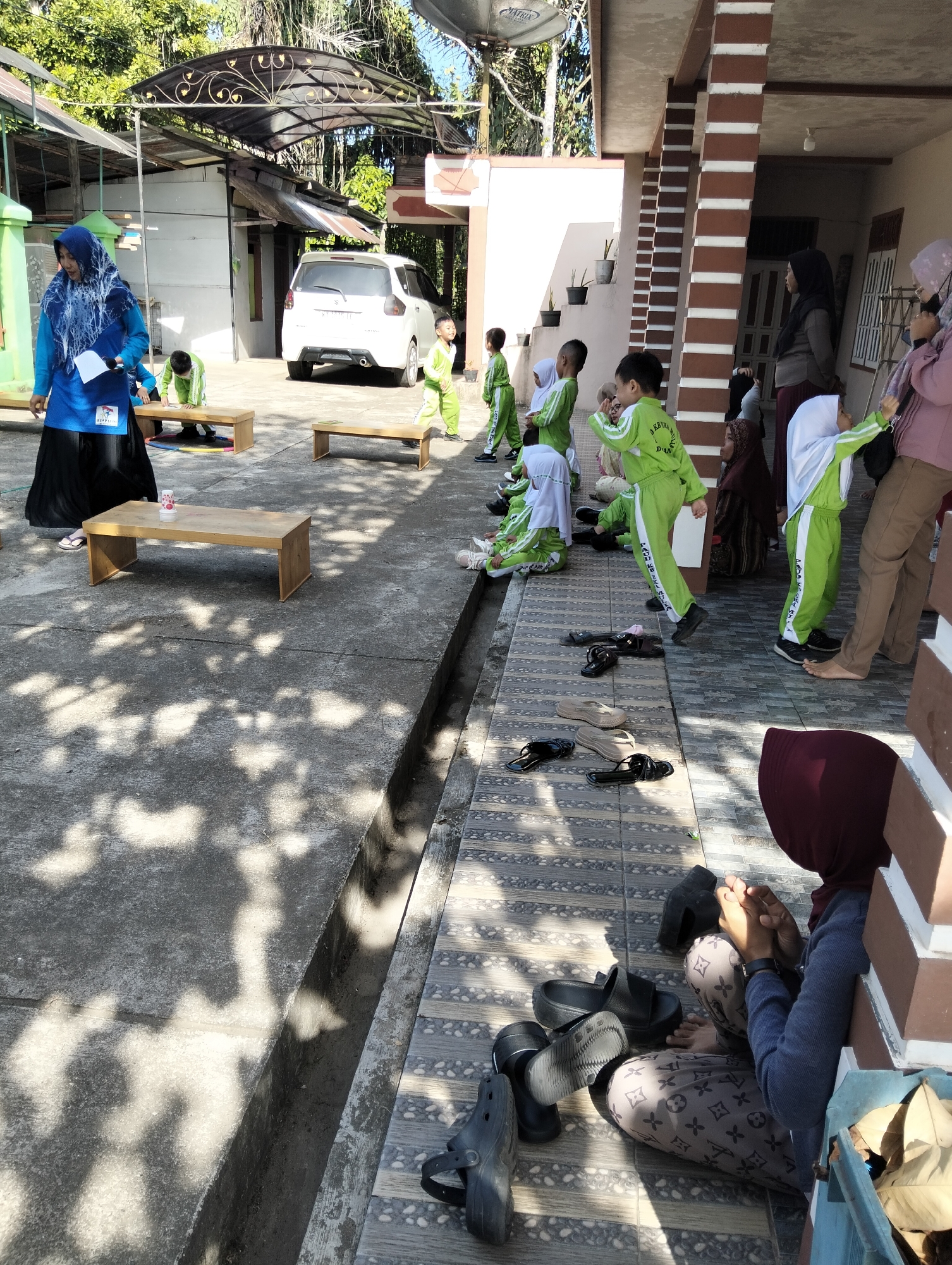 In Janju, Indonesia, a group of children, likely students, are gathered outdoors in what appears to be a school or community area. Several children are wearing bright green uniforms with white accents, and some are also wearing white headscarves. They are positioned in a line along a tiled walkway next to a building with a covered, open-air patio supported by brown and white striped columns. Some children are standing, while others are sitting on the ground or on low wooden benches. One child in a green uniform is captured in mid-motion, appearing to jump or dance. A woman in a blue and black outfit, possibly a teacher or guardian, stands to the left of the frame, holding a white paper. In the background, a white car is parked, and trees and other structures are visible, suggesting a semi-rural or suburban setting. The ground is a mix of concrete and tile, with shadows from overhead foliage dappling the surface. Scattered on the ground are various pairs of sandals and shoes. The scene is well-lit by sunlight.