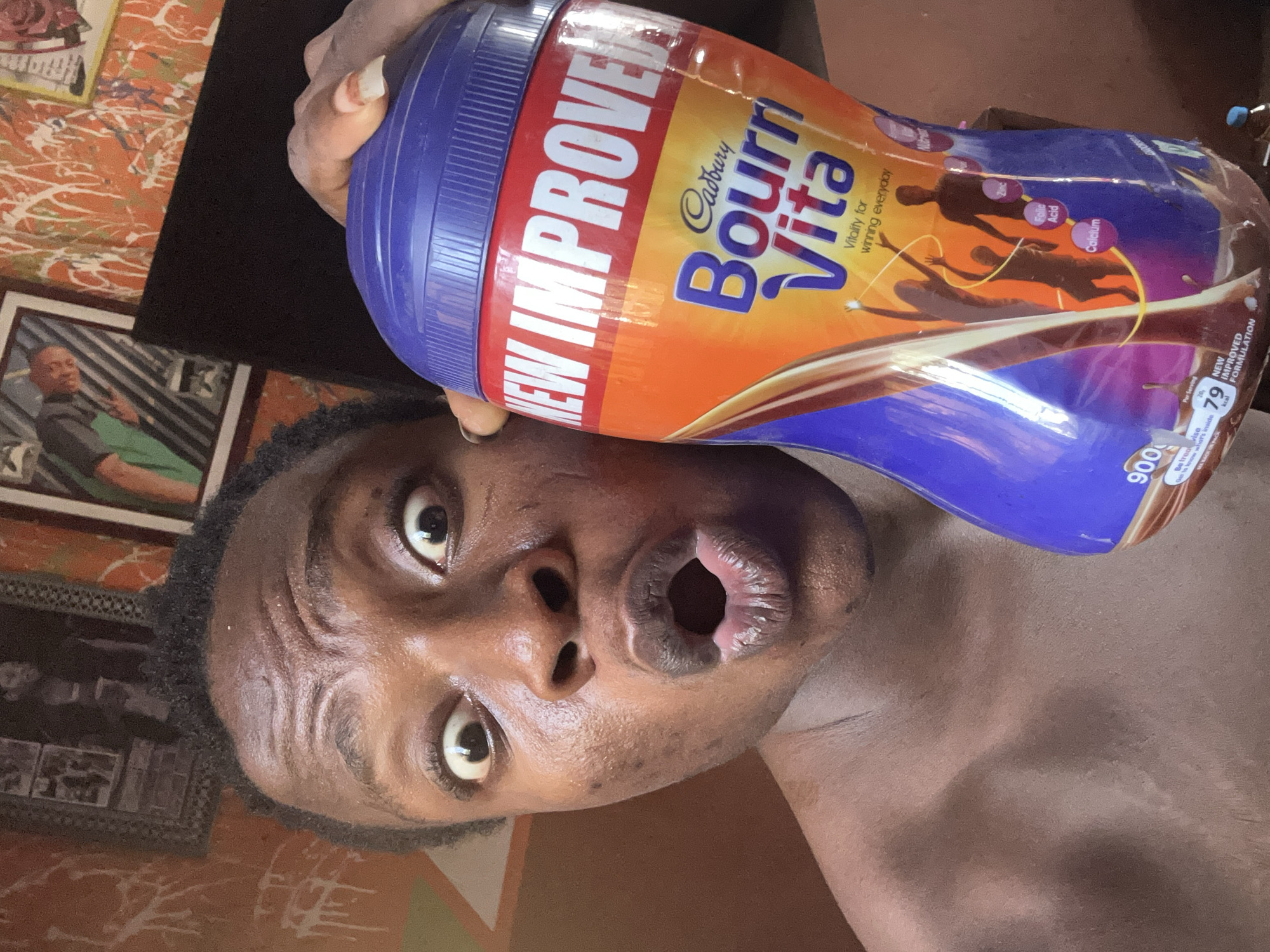 The image shows a dark-skinned man holding a container of Cadbury Bournvita, positioned close to his face. His eyes are wide and his mouth is open, suggesting a surprised or playful expression. The Bournvita container is blue and orange, with the words "NEW IMPROVED" and "Bournvita" prominently displayed. The container also mentions "Vitality for winning everyday" and lists folic acid and calcium. A picture frame can be seen in the background.