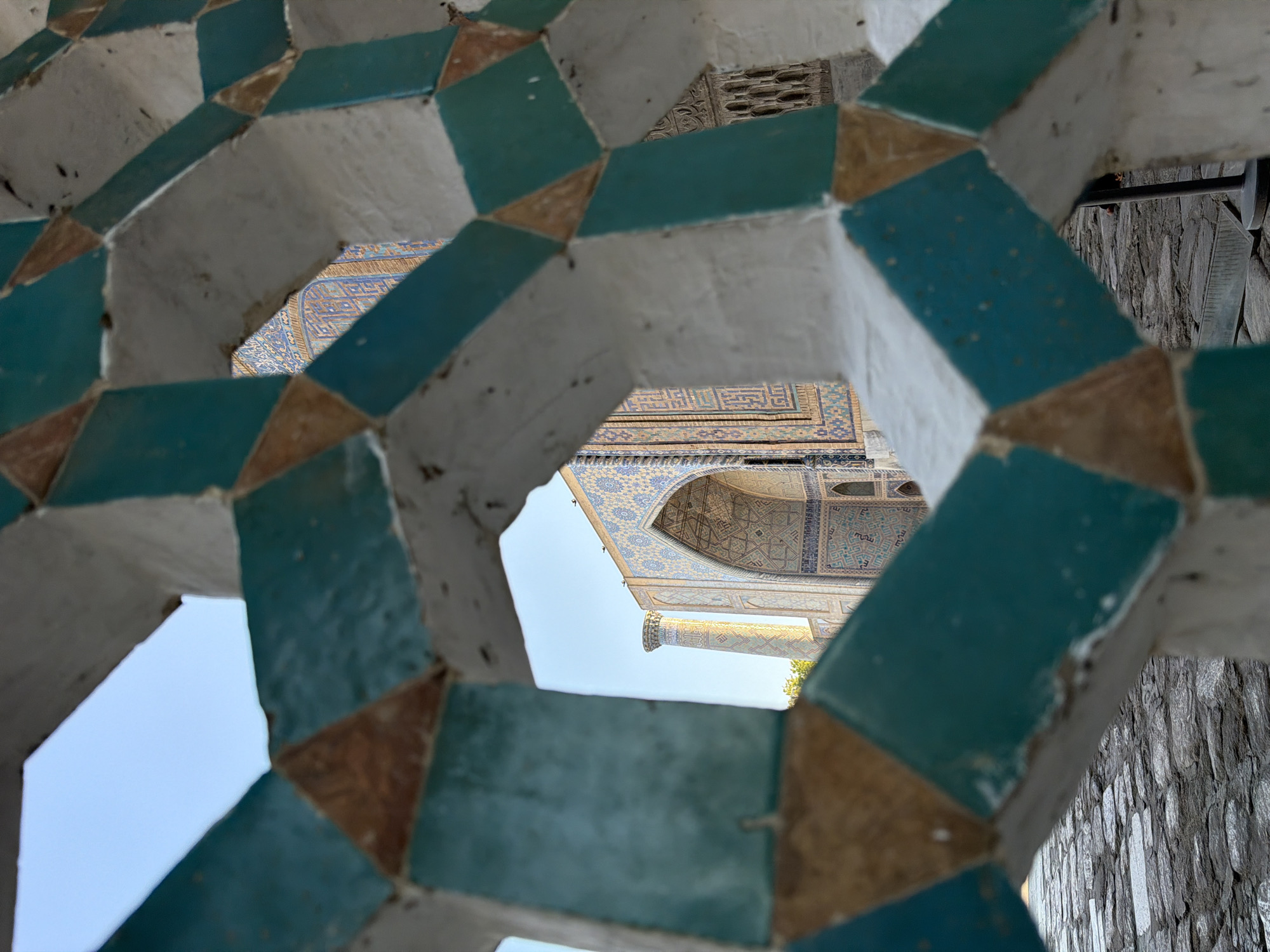 The image captures a detailed view of an ornate architectural structure in Samarkand City, Uzbekistan, likely part of a madrasah or mosque. In the foreground, a white, geometric lattice with turquoise and brown tiles forms a decorative screen, creating an aperture through which the background is viewed. This aperture reveals an archway and a minaret of a building adorned with intricate blue and yellow mosaic tiles. The stonework of the foreground path is visible at the bottom. The sky is a clear, pale blue.