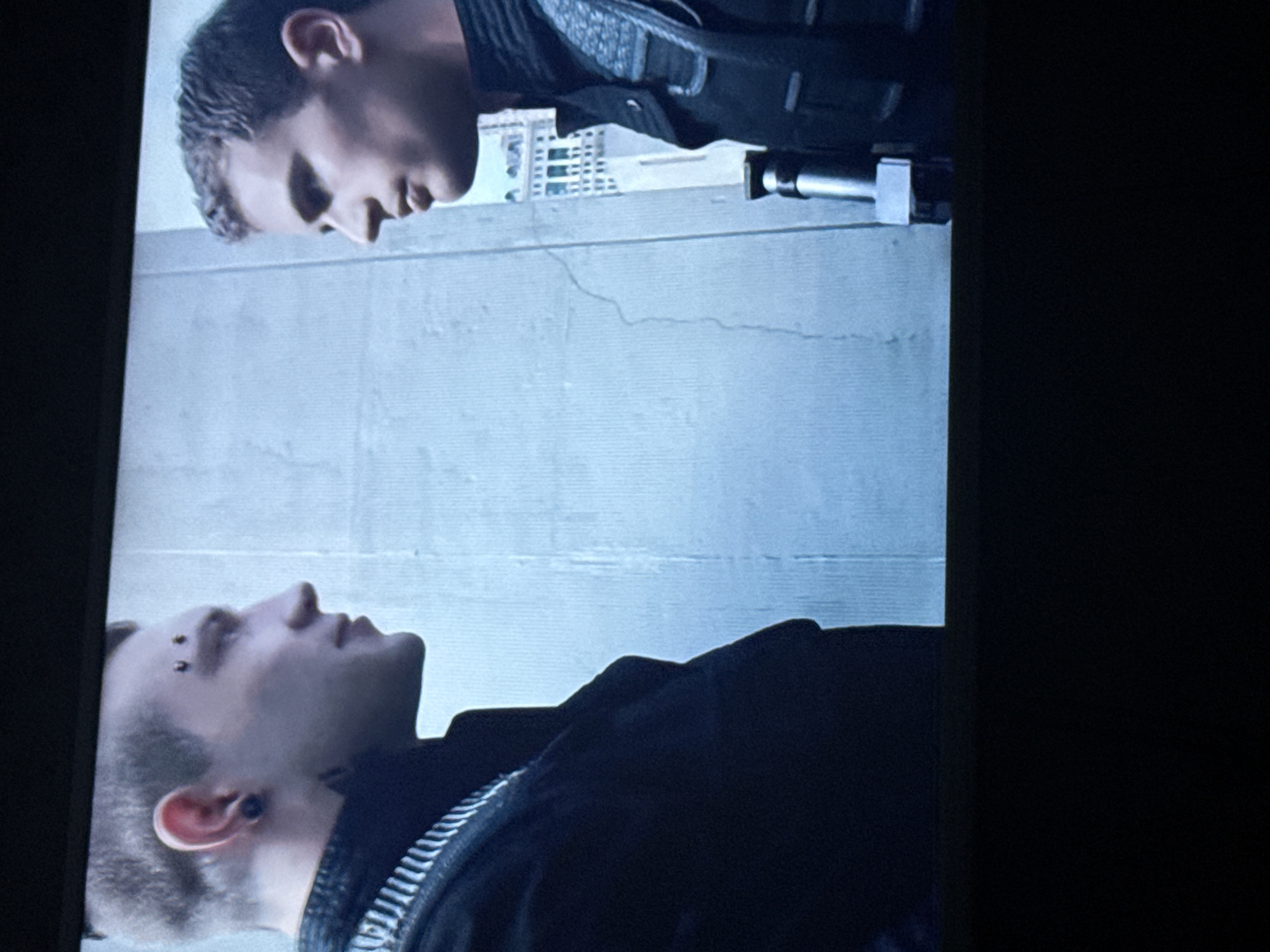 Two men are shown facing each other, seemingly indoors against a background of what appears to be a cityscape. The man at the top is wearing dark clothing with a gun is pointed at the man at the bottom who has eyebrow piercings and a dark tattoo. The building in the background does not appear to match architecture in Mytishchi, Russia.
