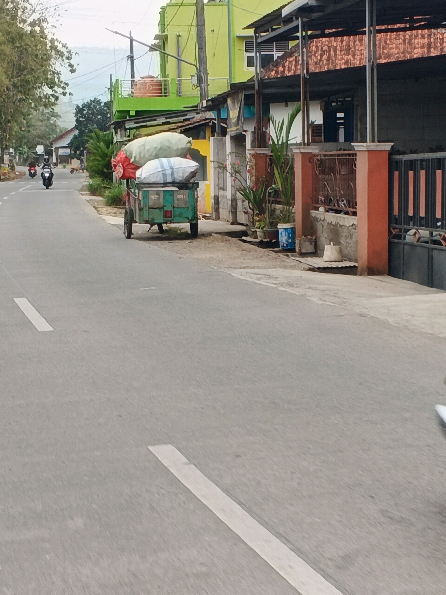 A street scene in Karangjengkol, Indonesia, shows a light green, motorized cart with two wheels, laden with large, white, and red sacks, parked on the right side of an asphalt road. The cart has a metal frame and a wooden body. Several motorcycles are visible on the road. On the right side of the road, there is a building with a green facade, a red tiled roof, and a concrete fence. The sky is overcast.