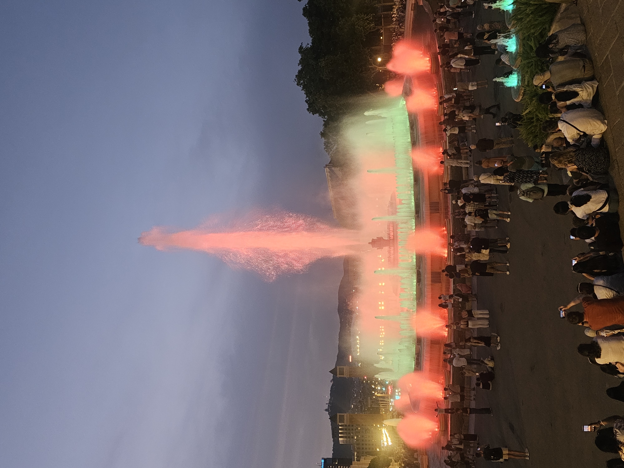 The image captures a lively evening scene at the Magic Fountain of Montjuïc in Barcelona, Spain. A large, illuminated fountain is the central focus, with jets of water lit in vibrant red and green hues, creating a dynamic display against the dusky sky. In the foreground and surrounding the fountain, a sizable crowd of people are gathered, many seated or standing, observing the spectacle. Several individuals are holding up smartphones, presumably to take photos or videos of the fountain. The background features the Barcelona skyline with buildings and distant hills, illuminated by city lights. The atmosphere appears festive and communal, with people of various ages enjoying the illuminated water show.
