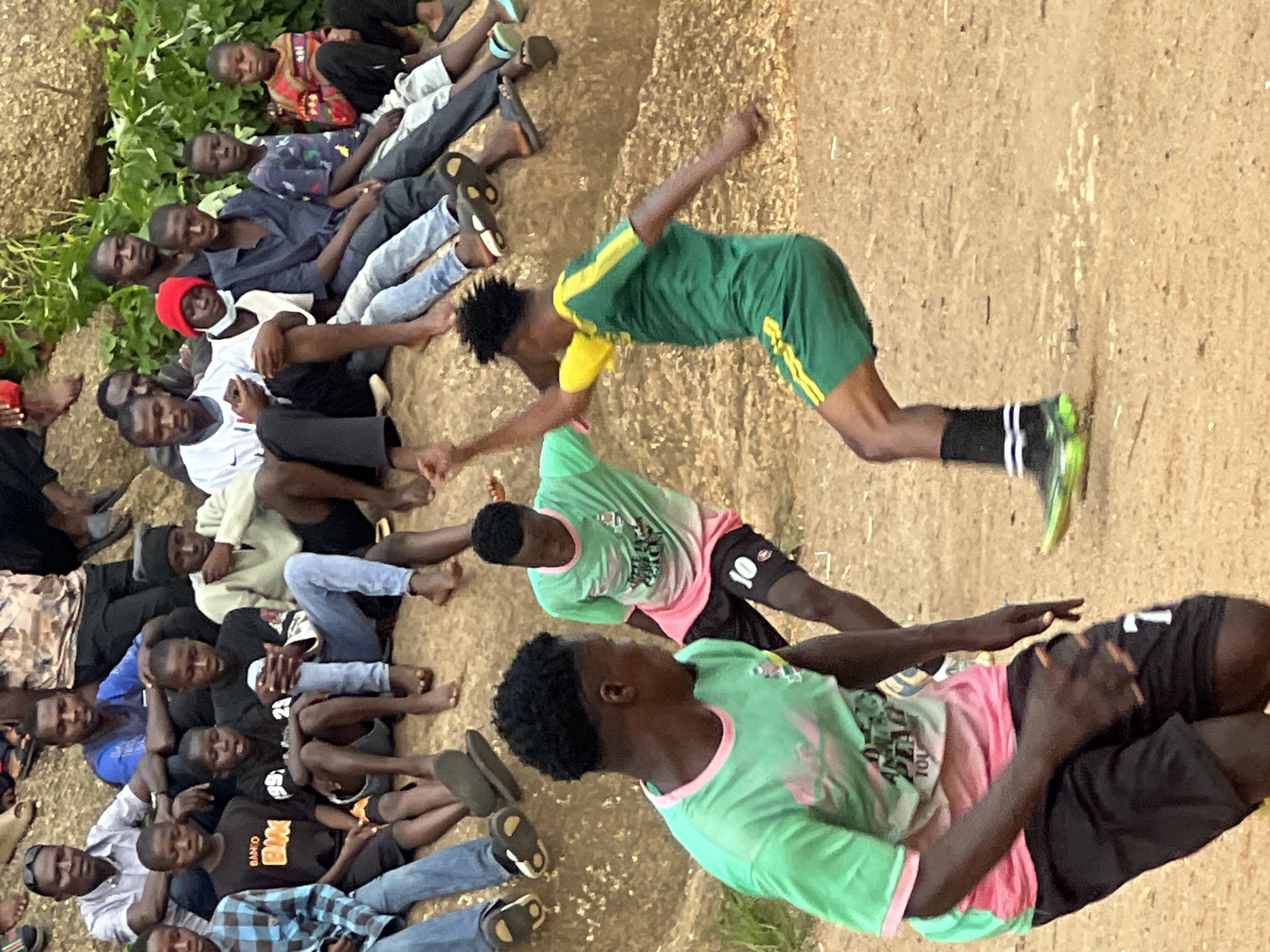 A group of individuals, predominantly young men and boys, are gathered outdoors on a rocky, uneven terrain. In the foreground, two individuals are actively engaged in a game, possibly soccer. One individual, wearing a green and yellow soccer uniform, is in motion, with one arm raised and bent. The other individual, wearing a pink and green jersey and black shorts with the number '7', is facing the first individual, with his left hand on his hip and his right arm bent. Several other people are seated on the rocks in the background, observing the activity. The environment appears to be dry and dusty, with sparse vegetation and large rocks. The scene suggests a casual gathering and recreational activity in an outdoor setting. The style of clothing and the presence of the soccer game suggest a location in a community in Jos, Nigeria, where such activities are common.