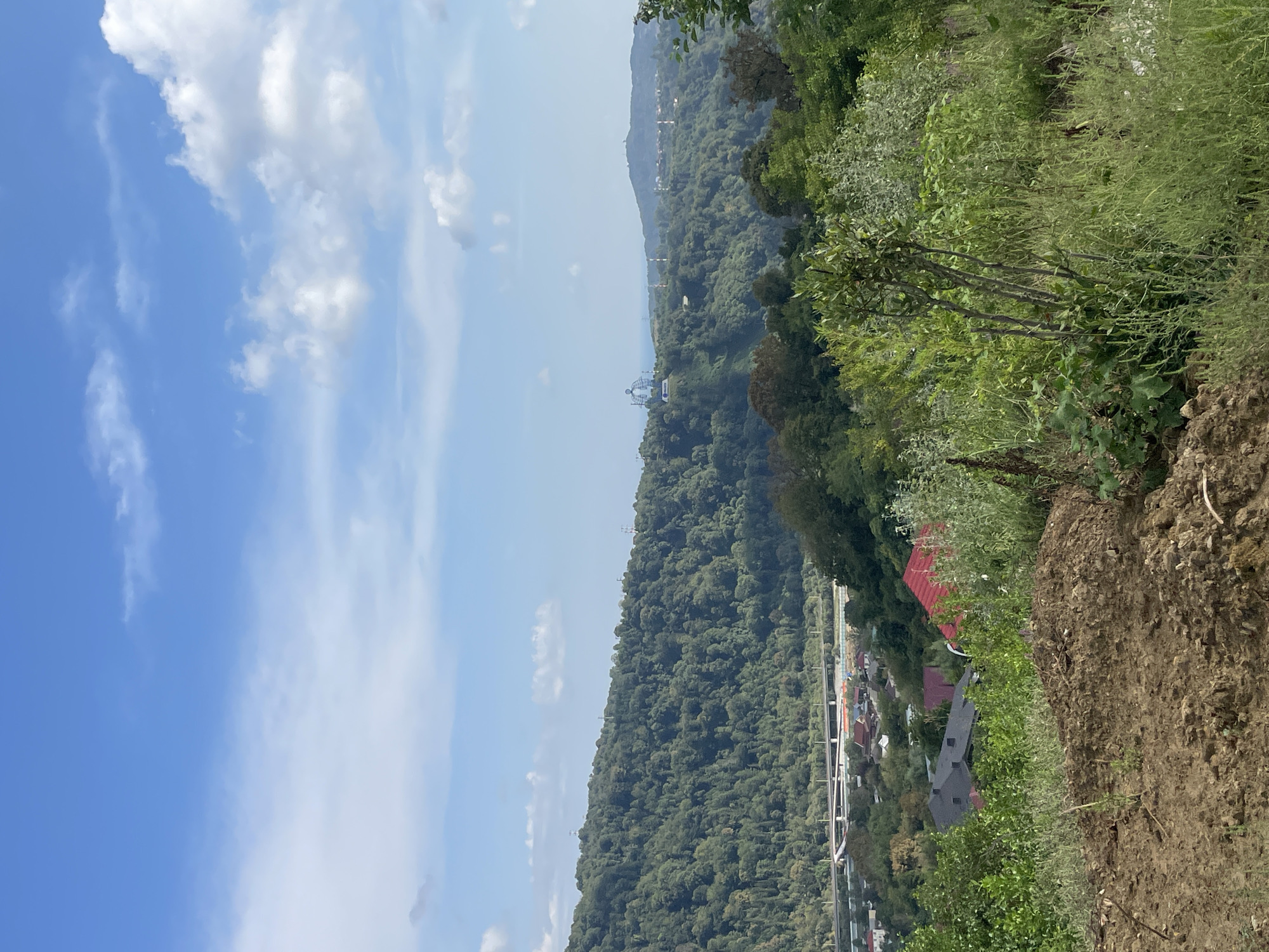 The image presents a panoramic view of a verdant landscape, likely captured from an elevated position, overlooking a settlement nestled amidst rolling hills. The foreground is dominated by a patch of disturbed earth, with a scattering of dry twigs and sparse vegetation.
Beyond this, a residential area is visible, characterized by a collection of houses with red and dark-tiled roofs, interspersed with trees and greenery. A bridge, possibly a highway overpass, spans across a lower area in the middle ground, suggesting a transportation route nearby.
The mid-ground and background are characterized by densely forested hills, covered in a rich tapestry of green foliage. In the distance, on one of the hilltops, a distinct white structure with a prominent dome is visible, hinting at a religious or landmark building. The sky above is a vibrant blue, dotted with fluffy white clouds, suggesting fair weather. The overall scene evokes a sense of rural tranquility and natural beauty, potentially within or near the city of Vysokoye, Russia, given the presence of Orthodox-style architecture. No people are visible in the frame.